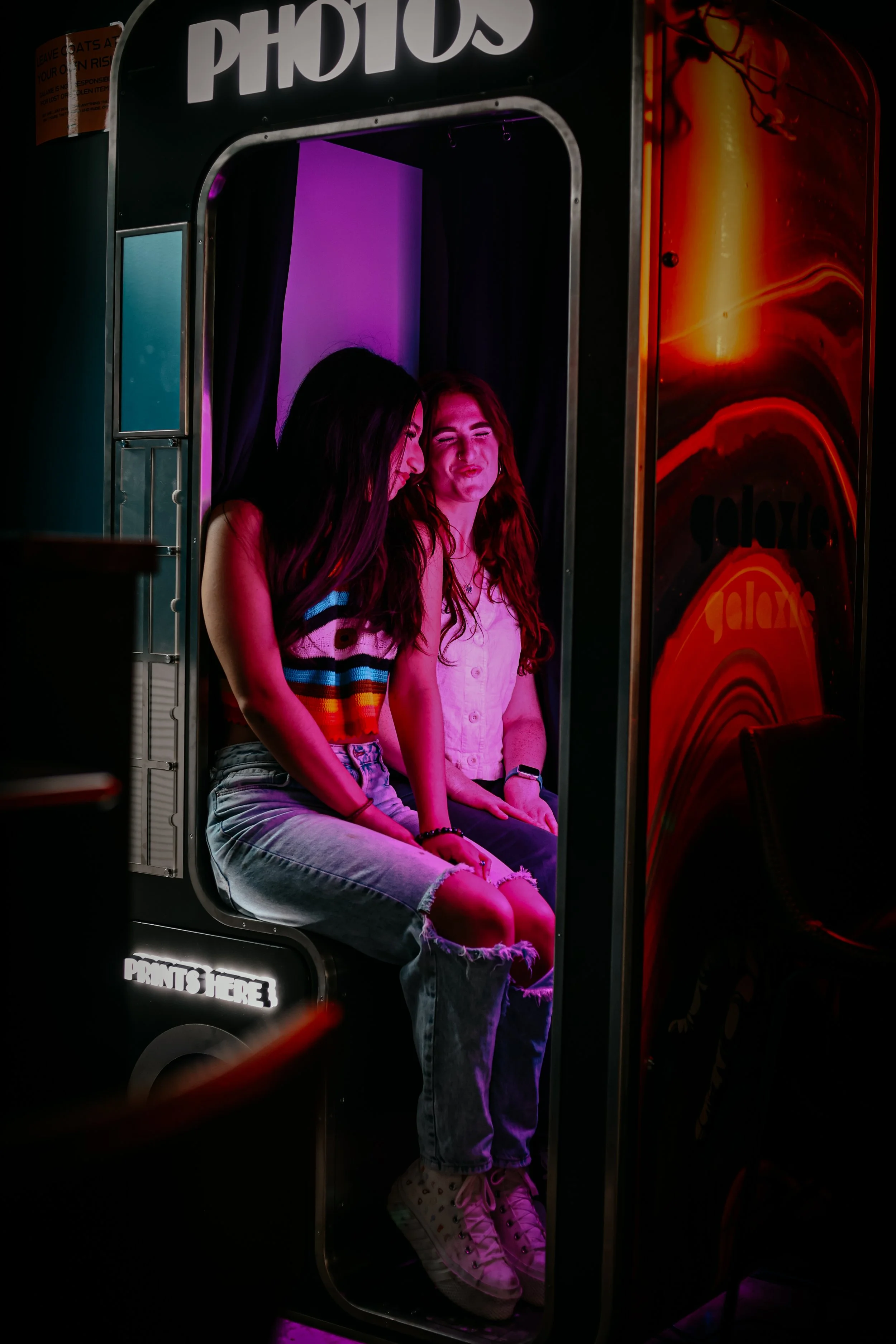 Two young women are sitting inside a photo booth with a dark background and purple lighting. One woman has long dark hair, a striped top, and distressed jeans, while the other has long red hair, a light pink top, and white sneakers. They appear to be laughing and having fun.
