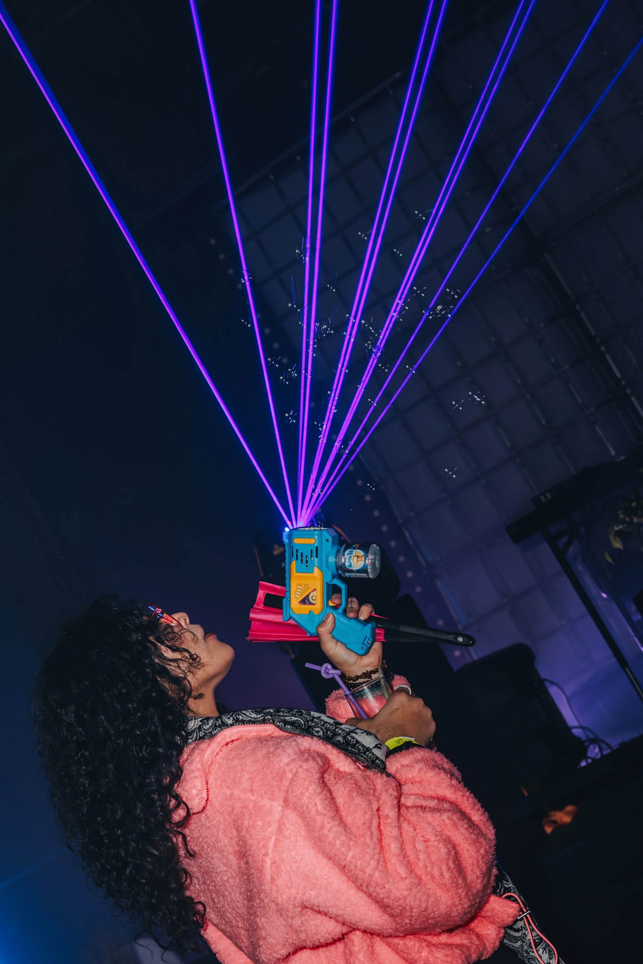 Woman holding a toy laser gun that emits blue and purple laser beams, wearing a pink fuzzy coat and multiple bracelets, at an indoor event with dark ceiling and lighting.