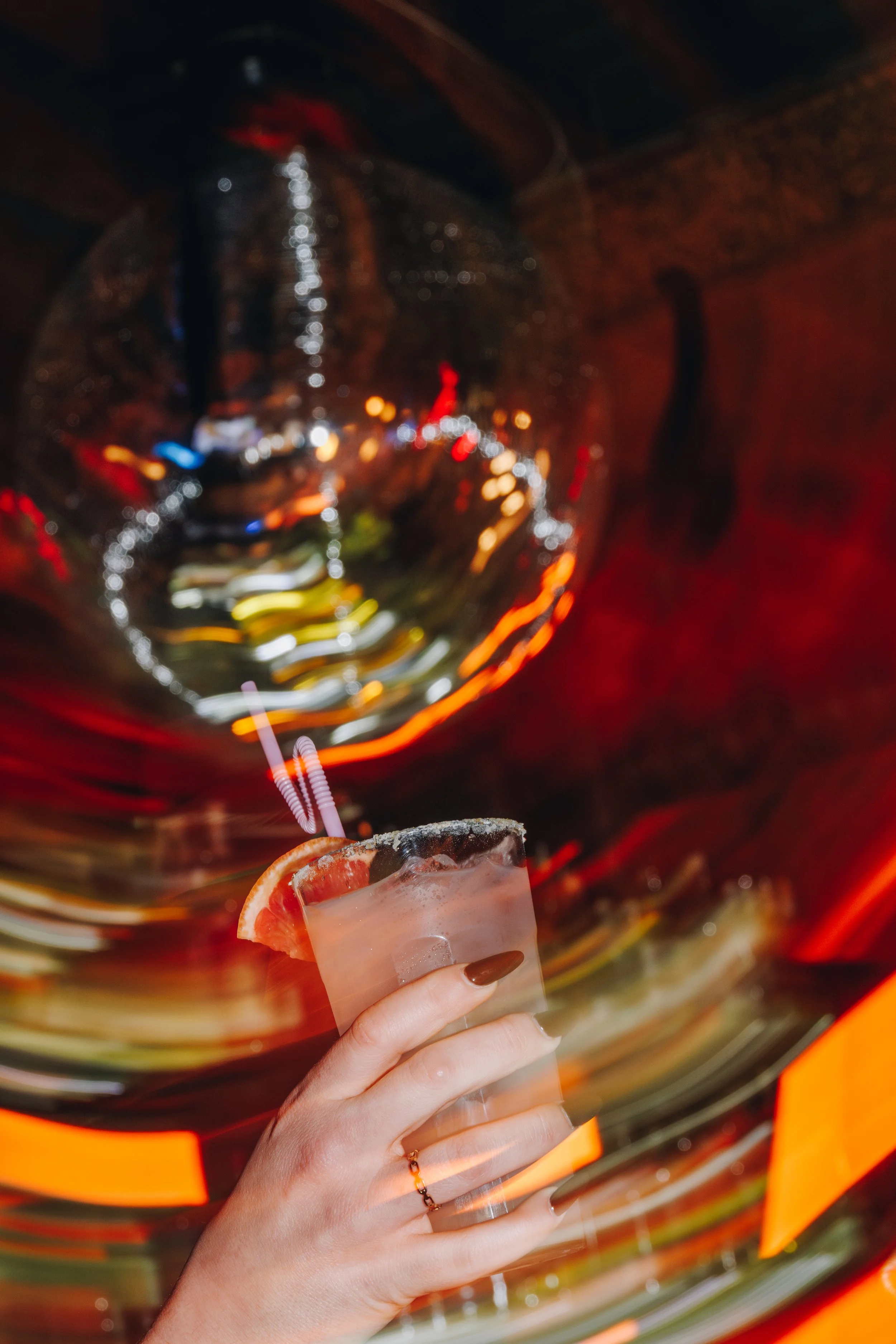 A person holding a cocktail glass with ice, a slice of grapefruit, and a straw, with colorful, blurred lights in the background.