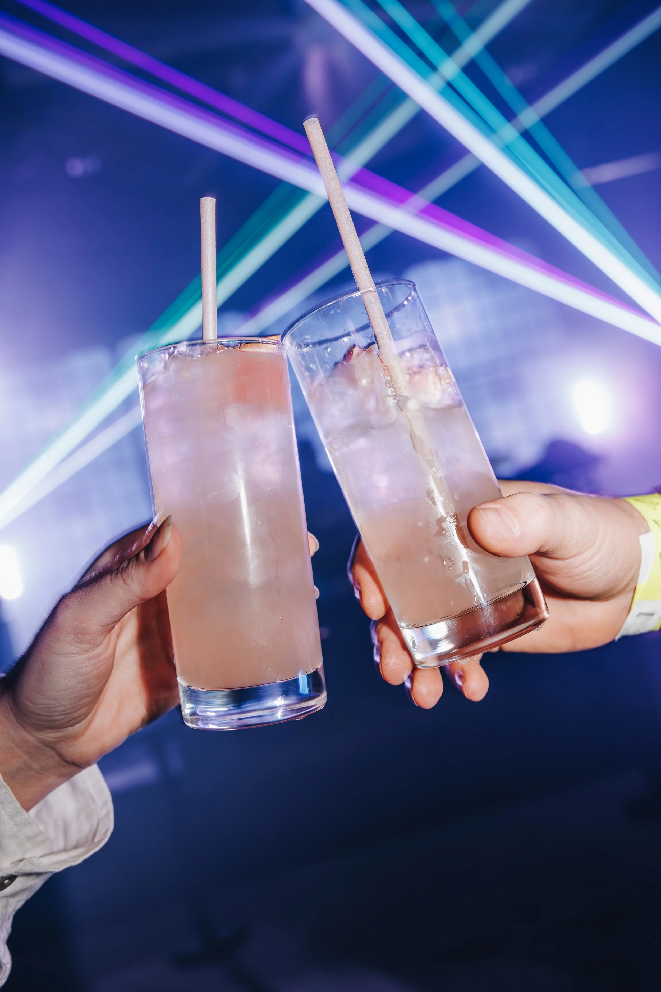 Three people clinking glasses filled with a light pink beverage, ice, and straws, against a backdrop of colorful laser lights.