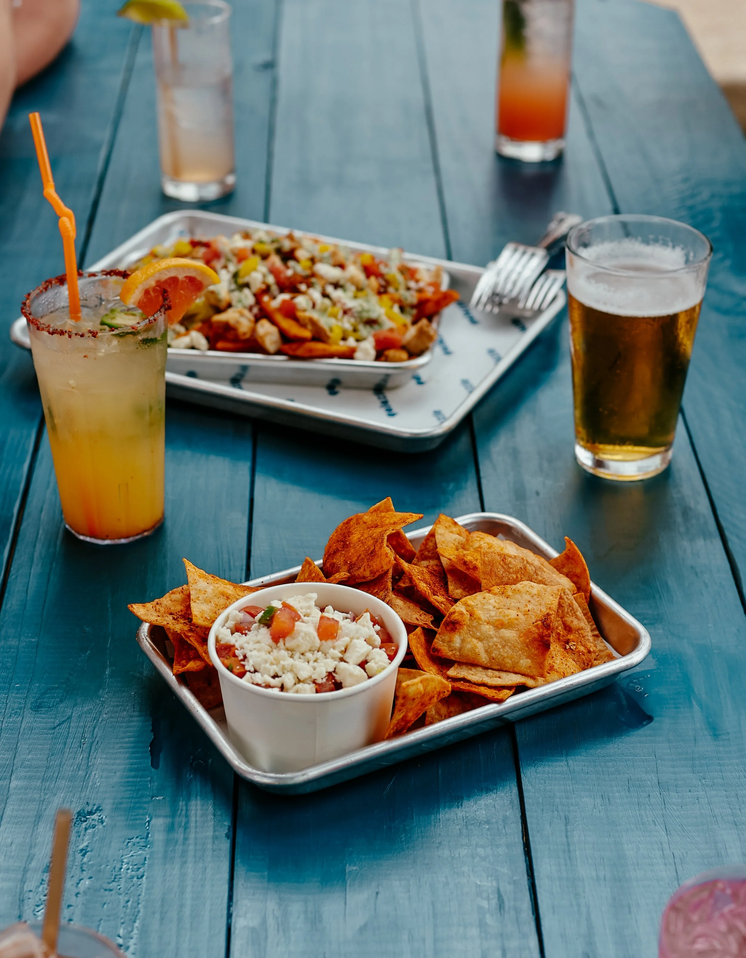 Tray of nachos with cheese, and a cup of cheese, with tortilla chips on a blue table alongside drinks, including a cocktail with a slice of orange and a green garnish, a glass of beer, and colorful cocktails in the background.