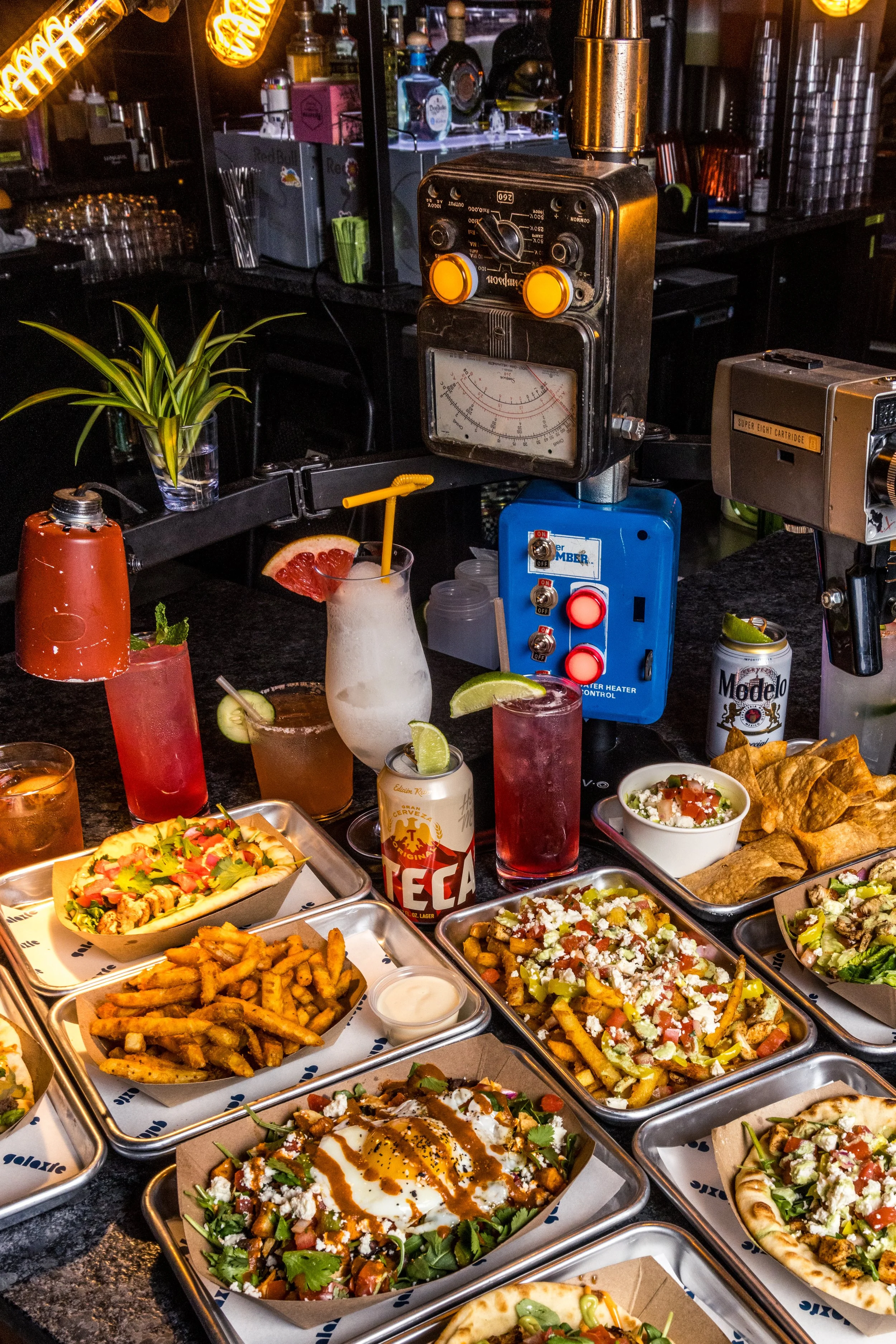 A bar counter with various drinks and cocktails, a potted plant, and dishes with loaded fries, salads, and pizza. There is a vintage soda fountain machine and bar equipment in the background.
