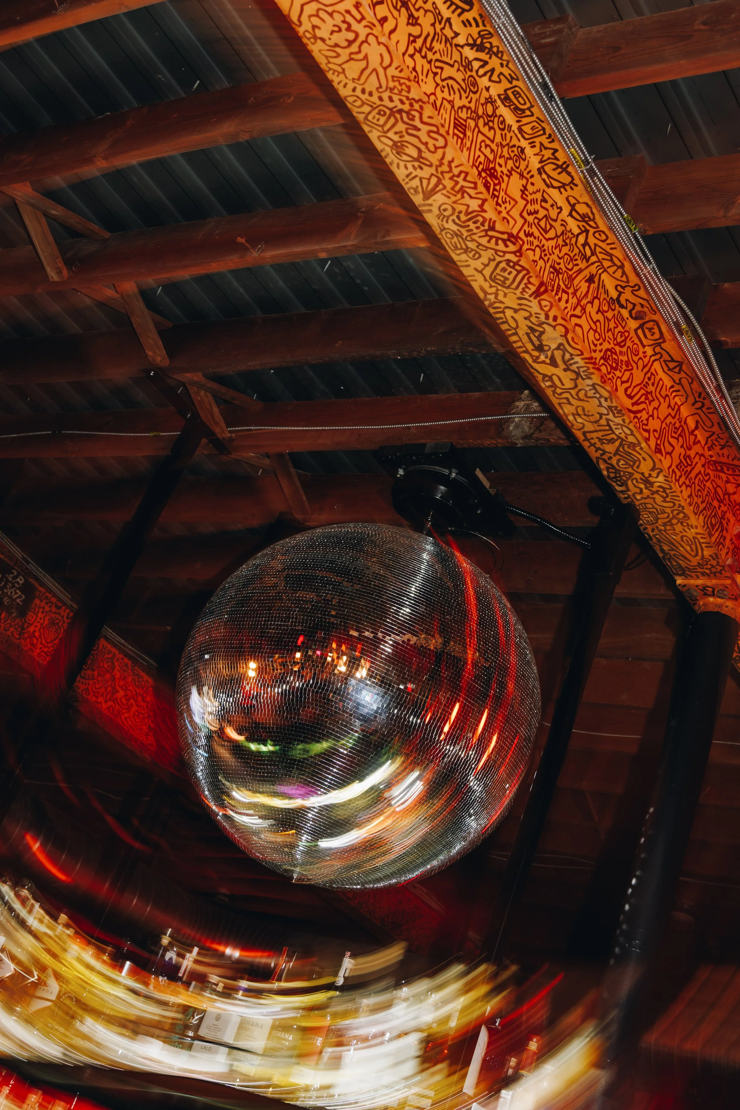 A reflective disco ball hanging from the ceiling, with streaks of light and motion blur creating a swirling pattern. The ceiling has exposed wooden beams and metal roofing, and part of a decorative cloth with abstract patterns is visible.