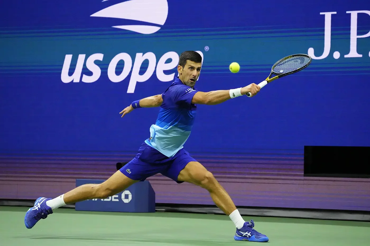 A male tennis player in a blue outfit lunges to hit a yellow tennis ball with his racket during a match at the US Open.