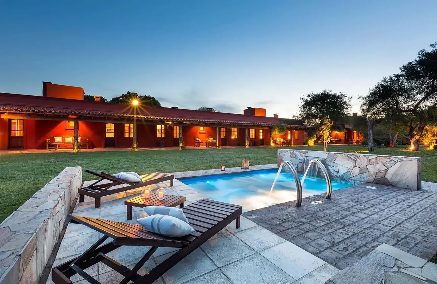 A peaceful outdoor scene at dusk featuring a lit swimming pool with jets, surrounded by lounge chairs, pillows, and candles, with a red building and trees in the background.