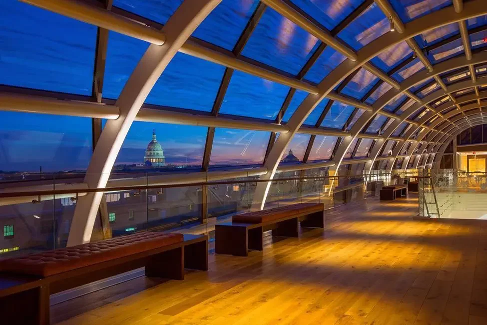 Night view from inside a modern, glass-enclosed observation deck with wooden benches, overlooking the U.S. Capitol building in Washington, D.C.