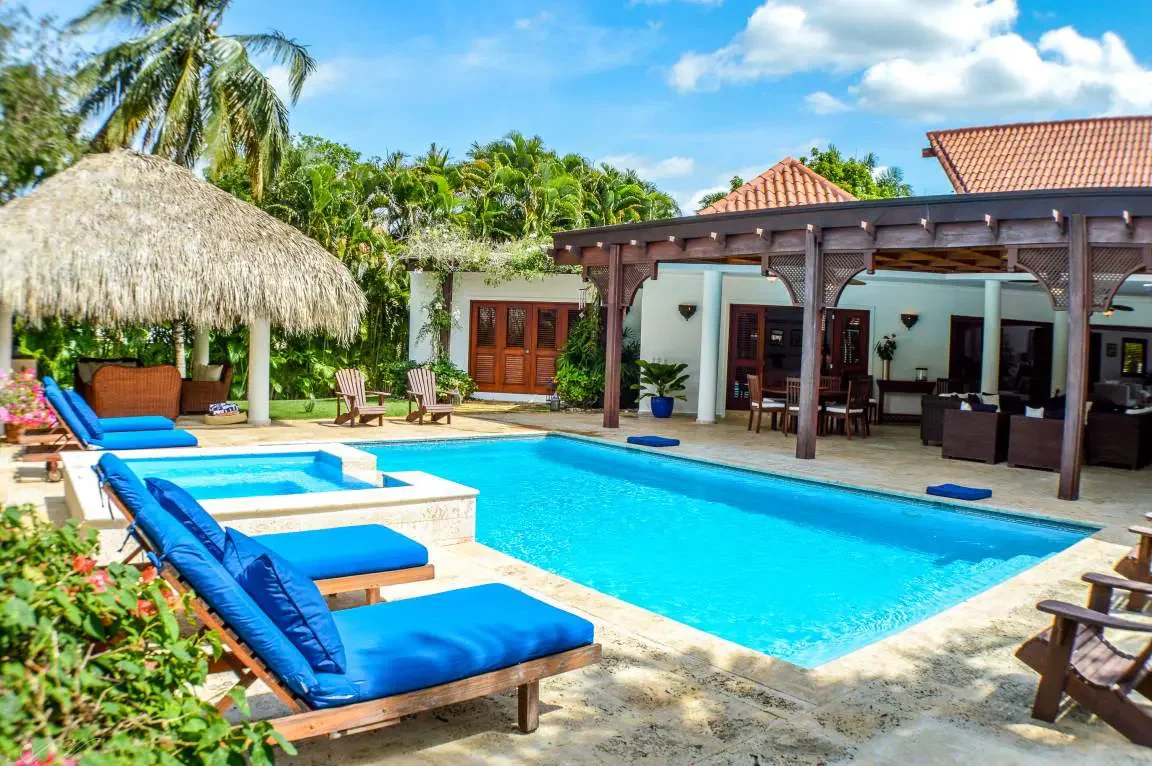 A backyard with a swimming pool, lounge chairs with blue cushions, a thatched-roof hut, and a covered patio area with outdoor furniture. There are green trees and a blue sky with some clouds in the background.