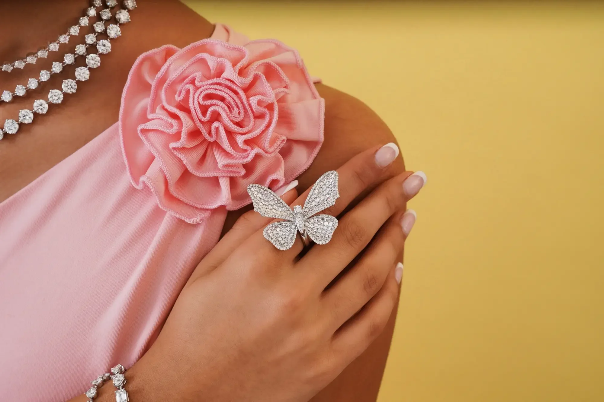 A woman wearing a necklace and a pink dress with a large pink ruffled flower on the shoulder, holding a silver butterfly ring. She has a diamond bracelet and a diamond ring on her fingers, with a yellow background.