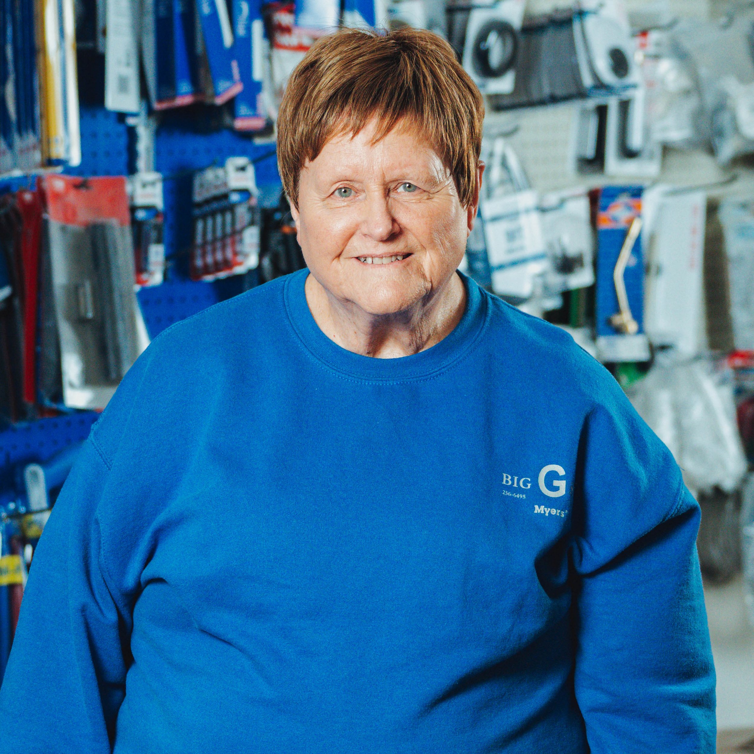 An elderly woman in a blue work shirt standing in a hardware store aisle surrounded by tools and hardware supplies.