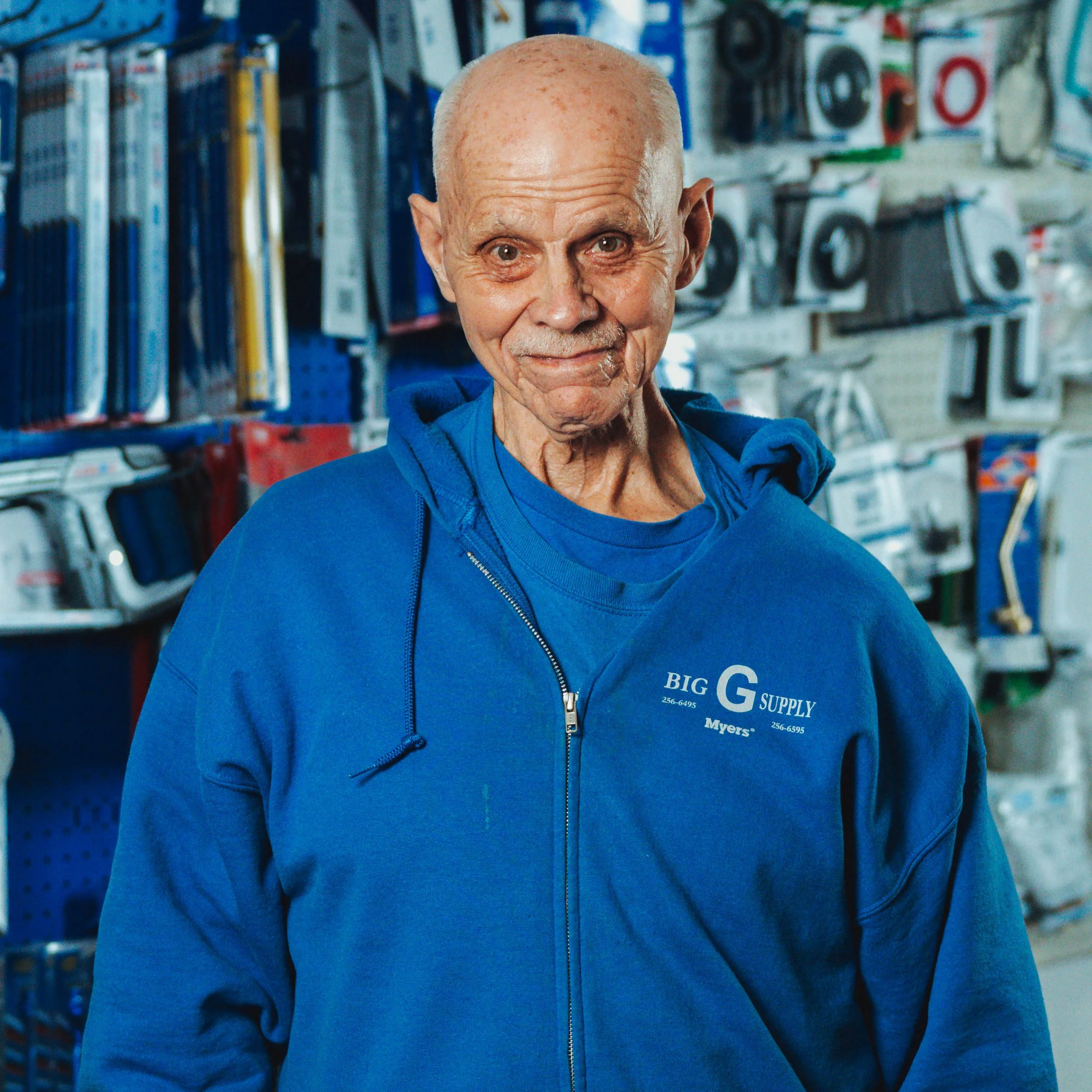 An elderly man with a bald head and light skin, wearing a blue hoodie that says 'Big G Supply' and 'Myers' on it, standing inside a store with various household items on shelves in the background.