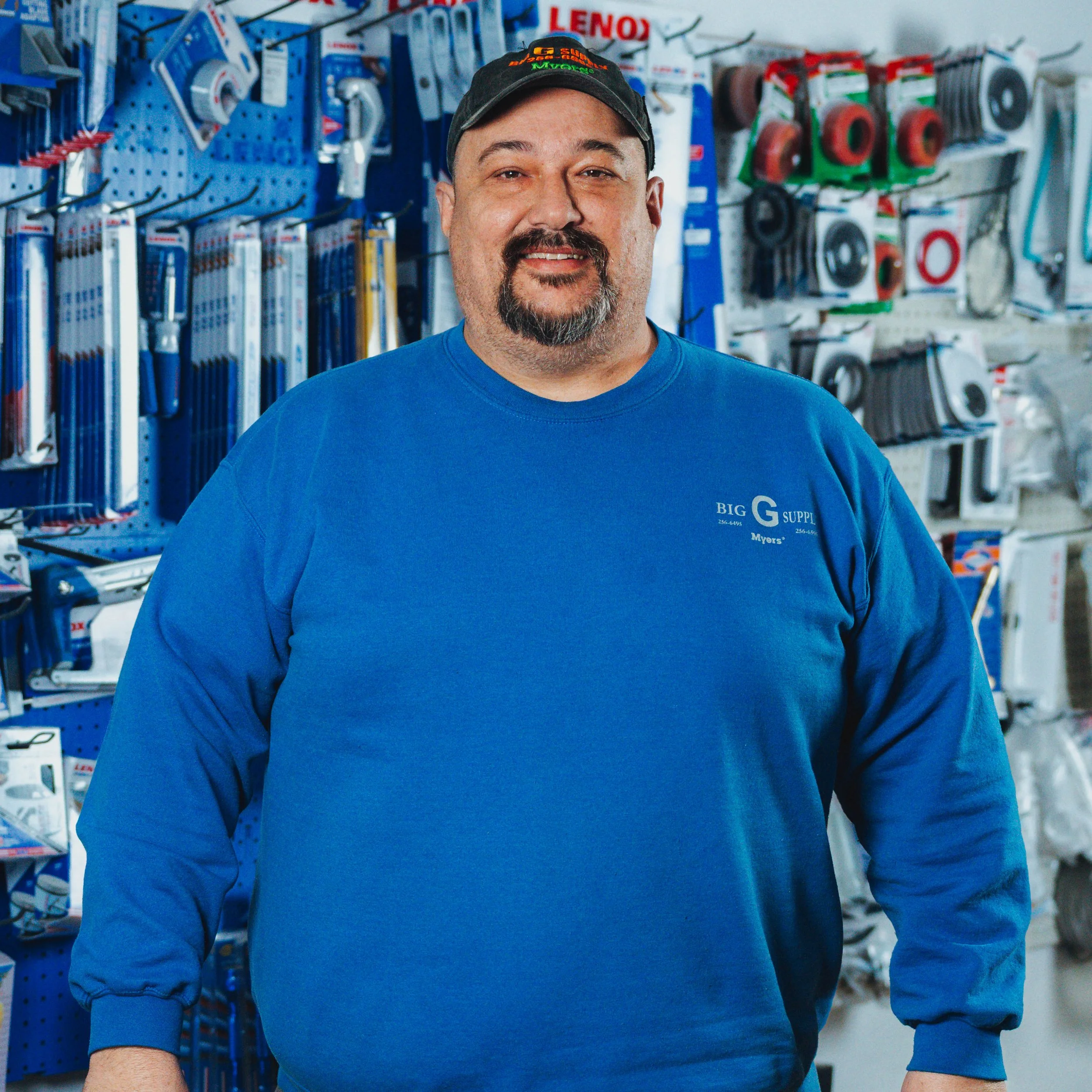 A man with a beard and mustache wearing a blue long-sleeve work shirt and a black baseball cap, standing inside a hardware store with tools and supplies on the shelves behind him.