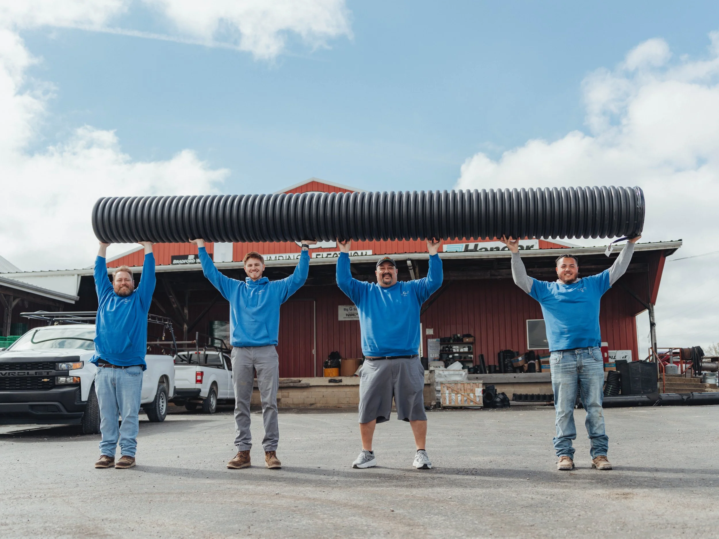 Four men in blue shirts standing outdoors in front of a building, holding a large black pipe above their heads.