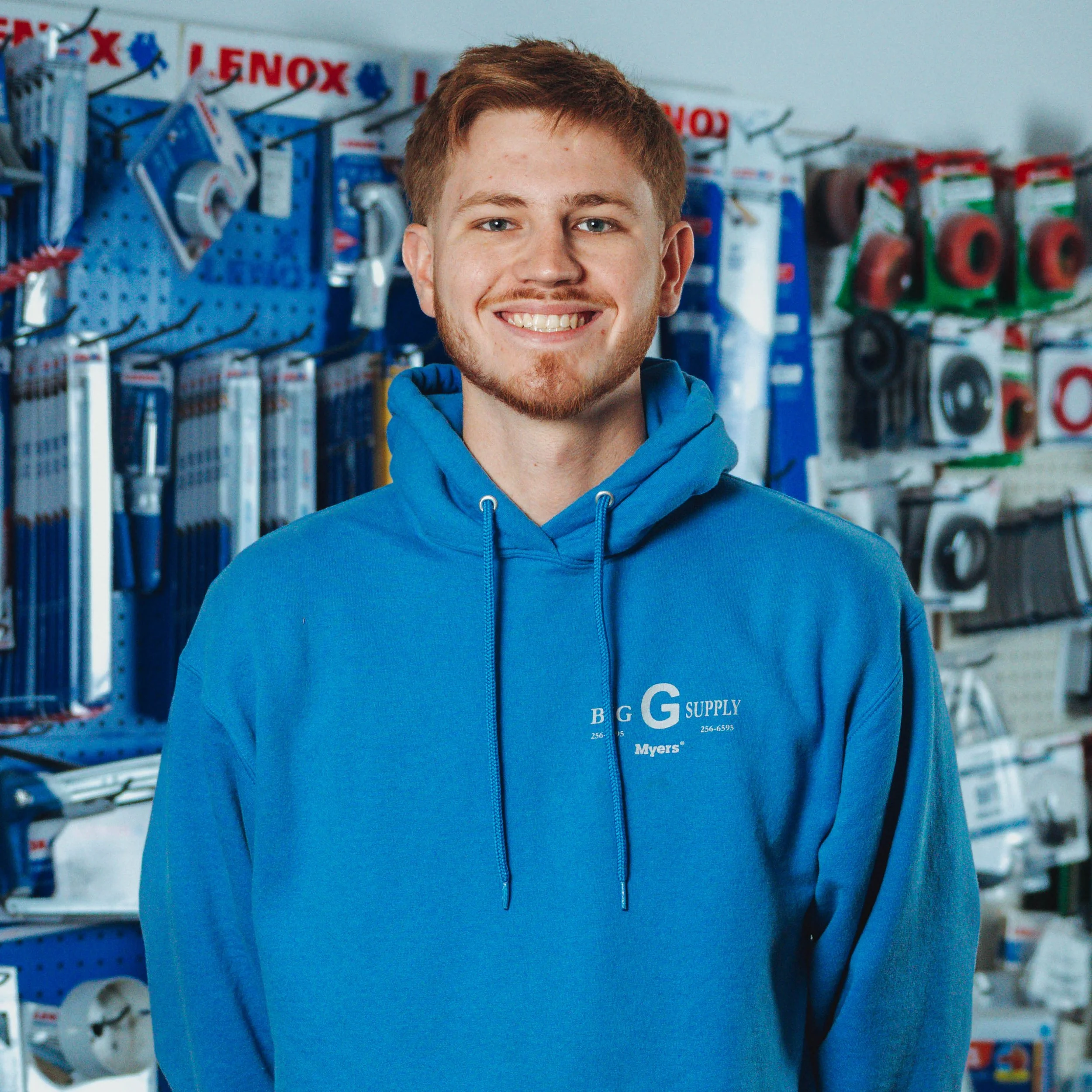 A young man with short red hair and a beard, smiling, wearing a blue hoodie with 'Big G Supply' logo, standing inside a hardware store with assorted tools and supplies on the shelves behind him.
