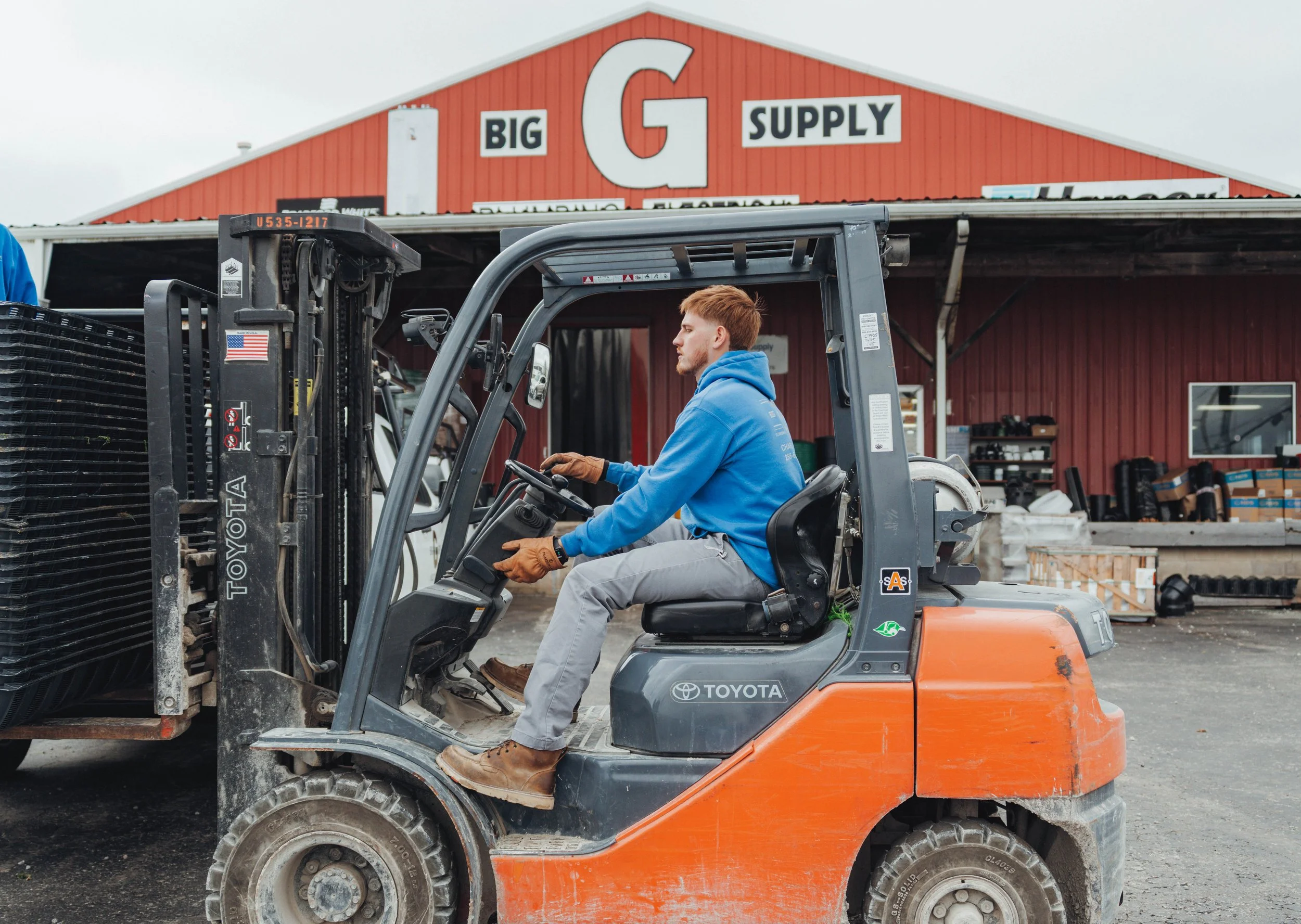 Young man in blue hoodie sitting on the control seat of an orange Toyota forklift, operating it in front of a red building with a sign that says "Big G Supply."