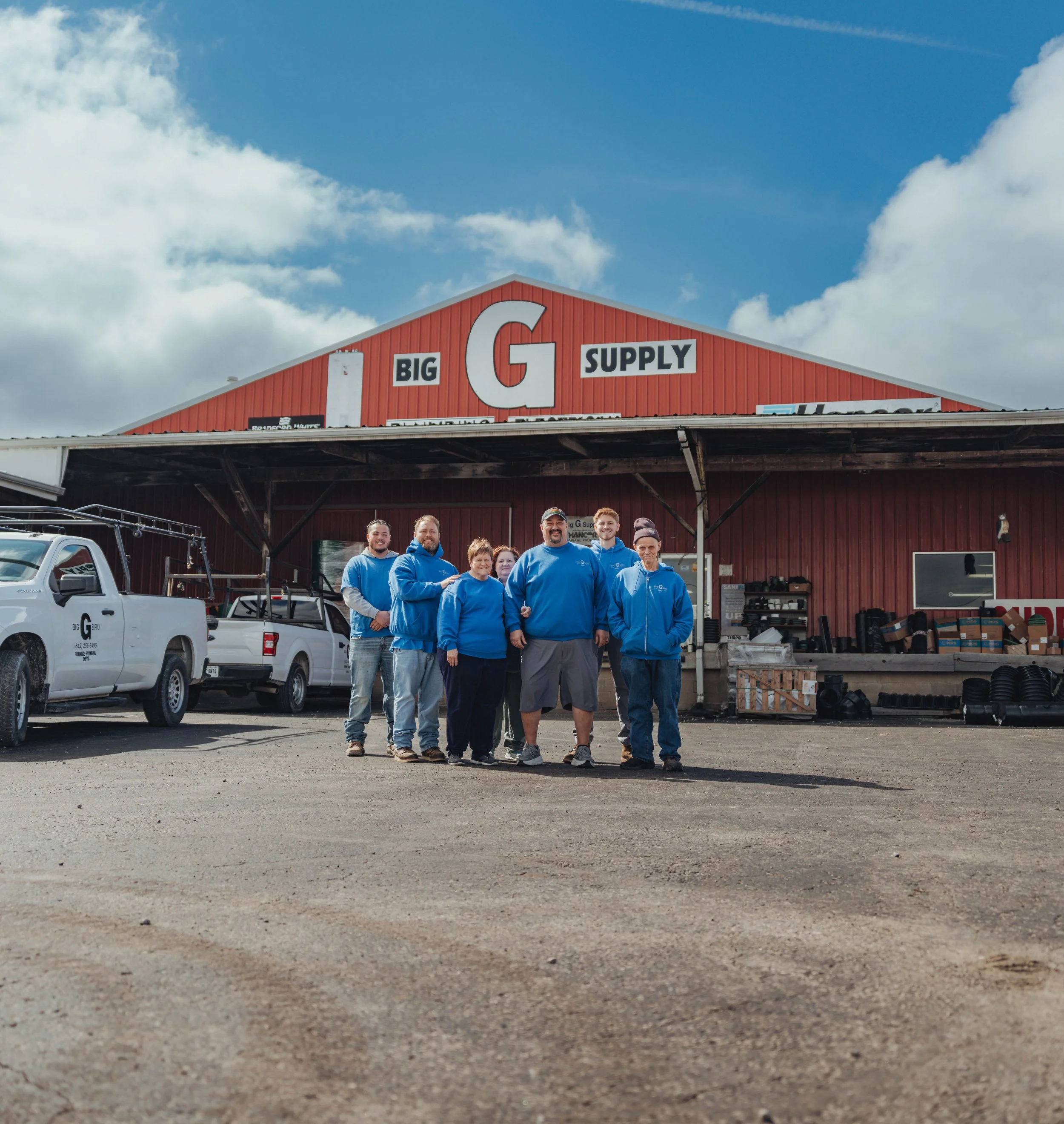Group of seven employees standing outside Big G Supply store, a red building with large sign, and trucks parked nearby, under partly cloudy sky.