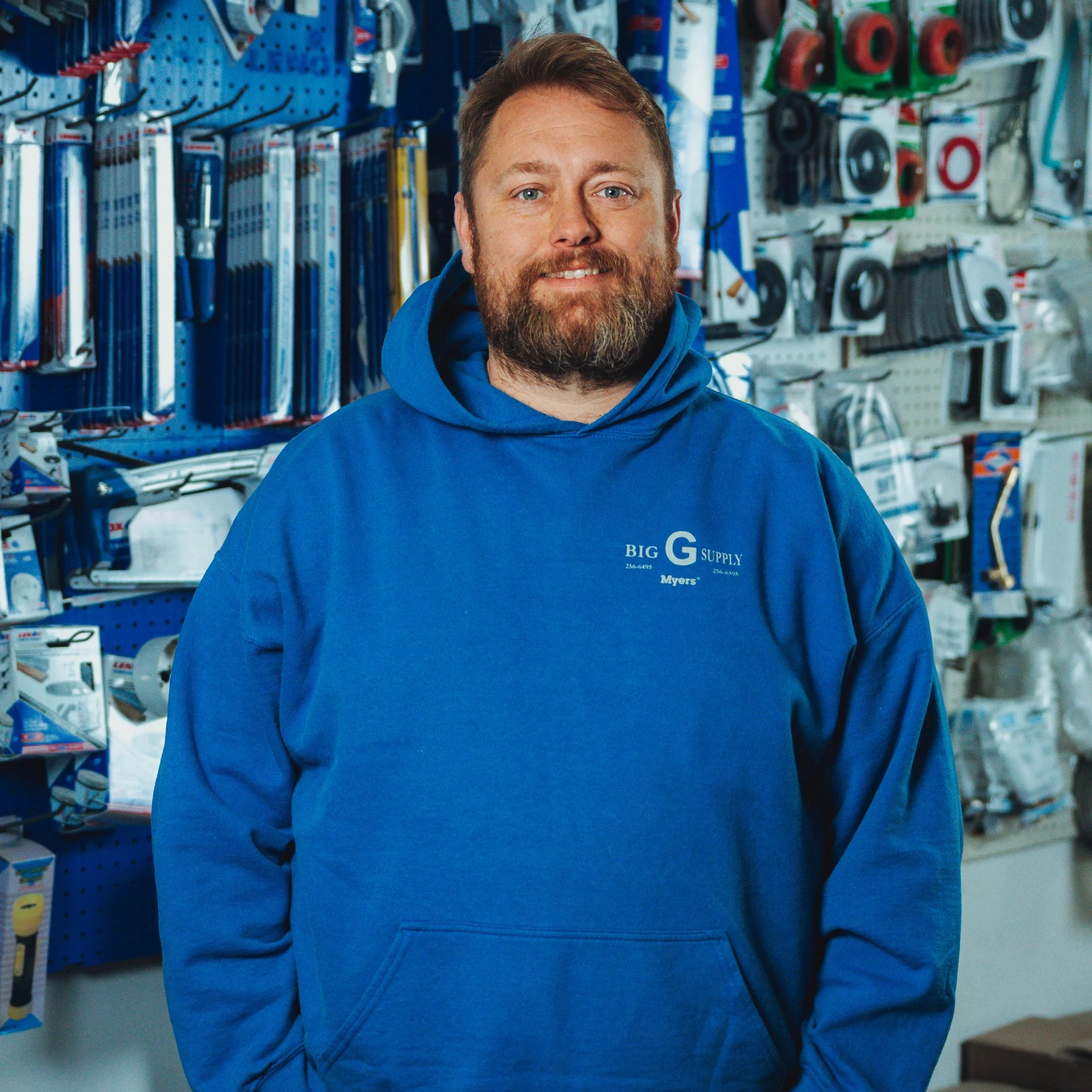 A man with a beard and light brown hair, smiling, wearing a blue hoodie that says "Big G Supply". He is standing inside a hardware store with various tools and supplies hanging on the wall behind him.