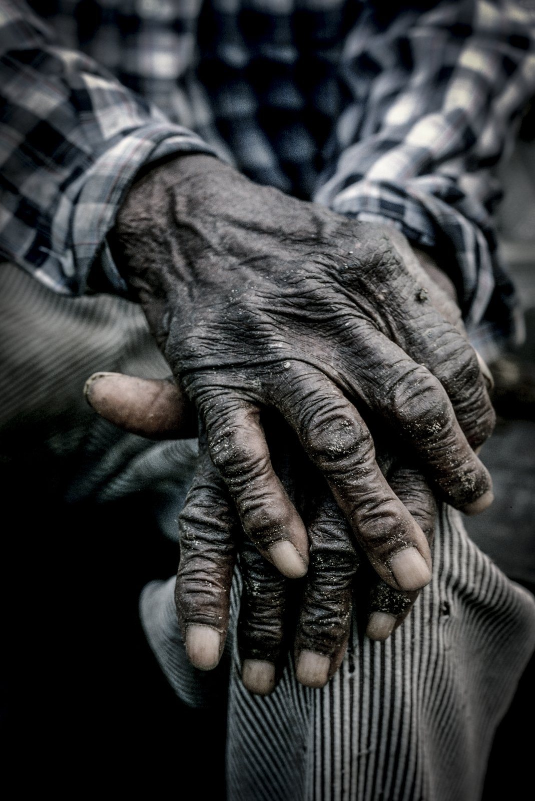 Historical image from 1987 of an older Gullah Geechee person's hands on top of on another, resting on their knee. They are wearing striped pants and a black and white flannel shirt.