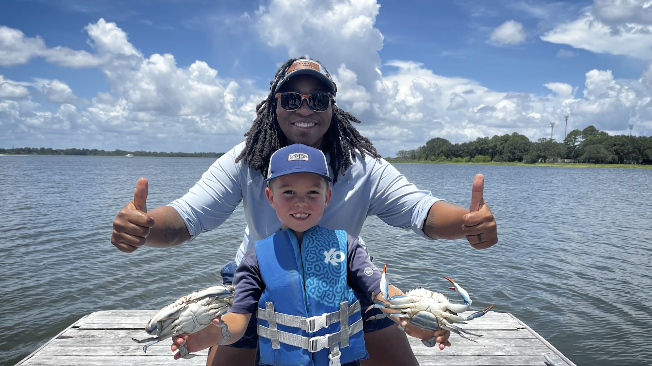 Tia Clark and a young boy on a boat on the water. Tia Clark has both thumbs up, and the boy is wearing a life jacket and a baseball cap and is holding two crabs.