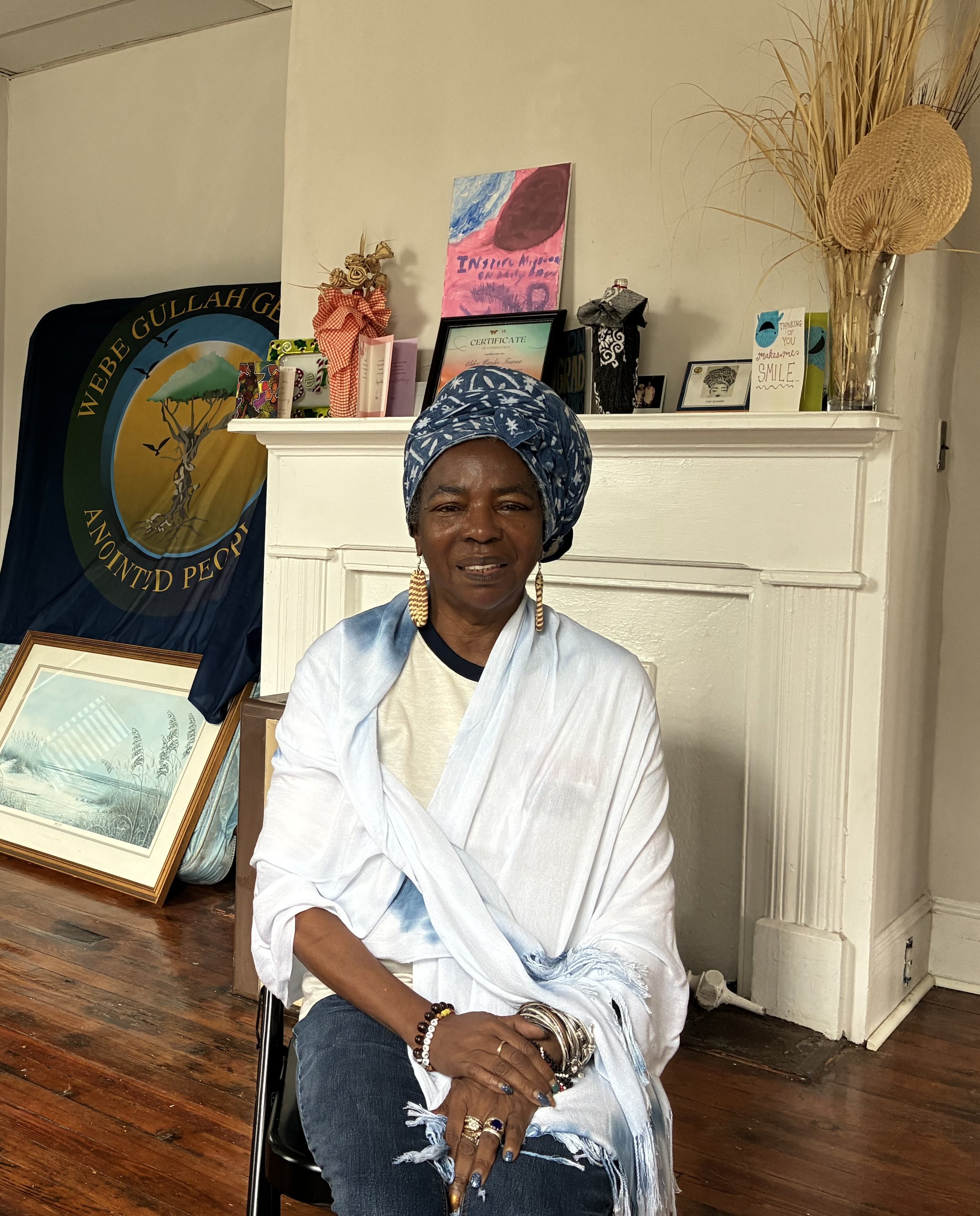 Elder Carlie Town smiling, sitting inside on a chair in front of a closed fireplace with cards, art, and foliage on the mantle. In the back against the wall is the flag for the Gullah Geechee Nation.