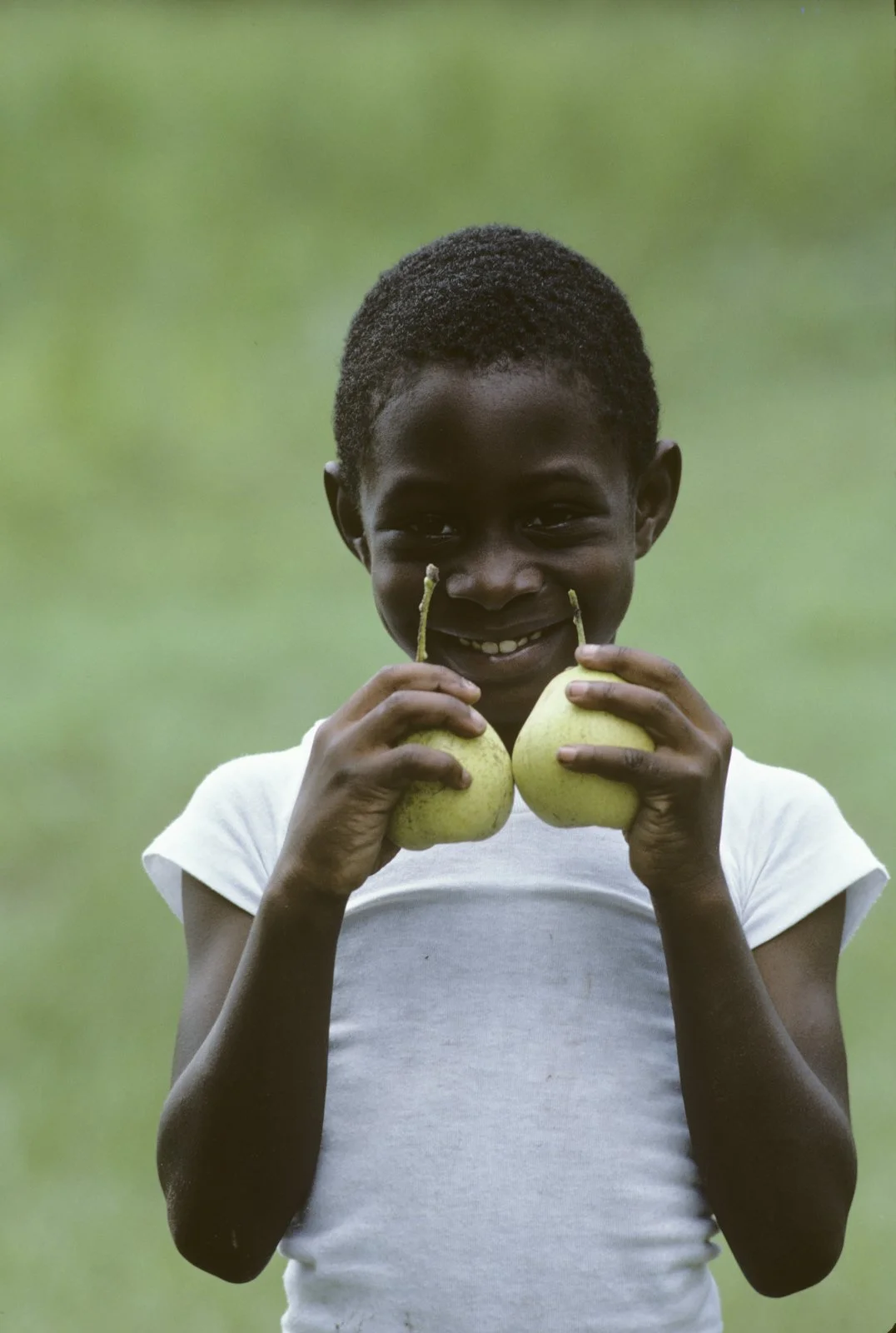 Historical image from 1987 of a small Gullah Geechee child wearing a white shirt, smiling, and holding two small pears.