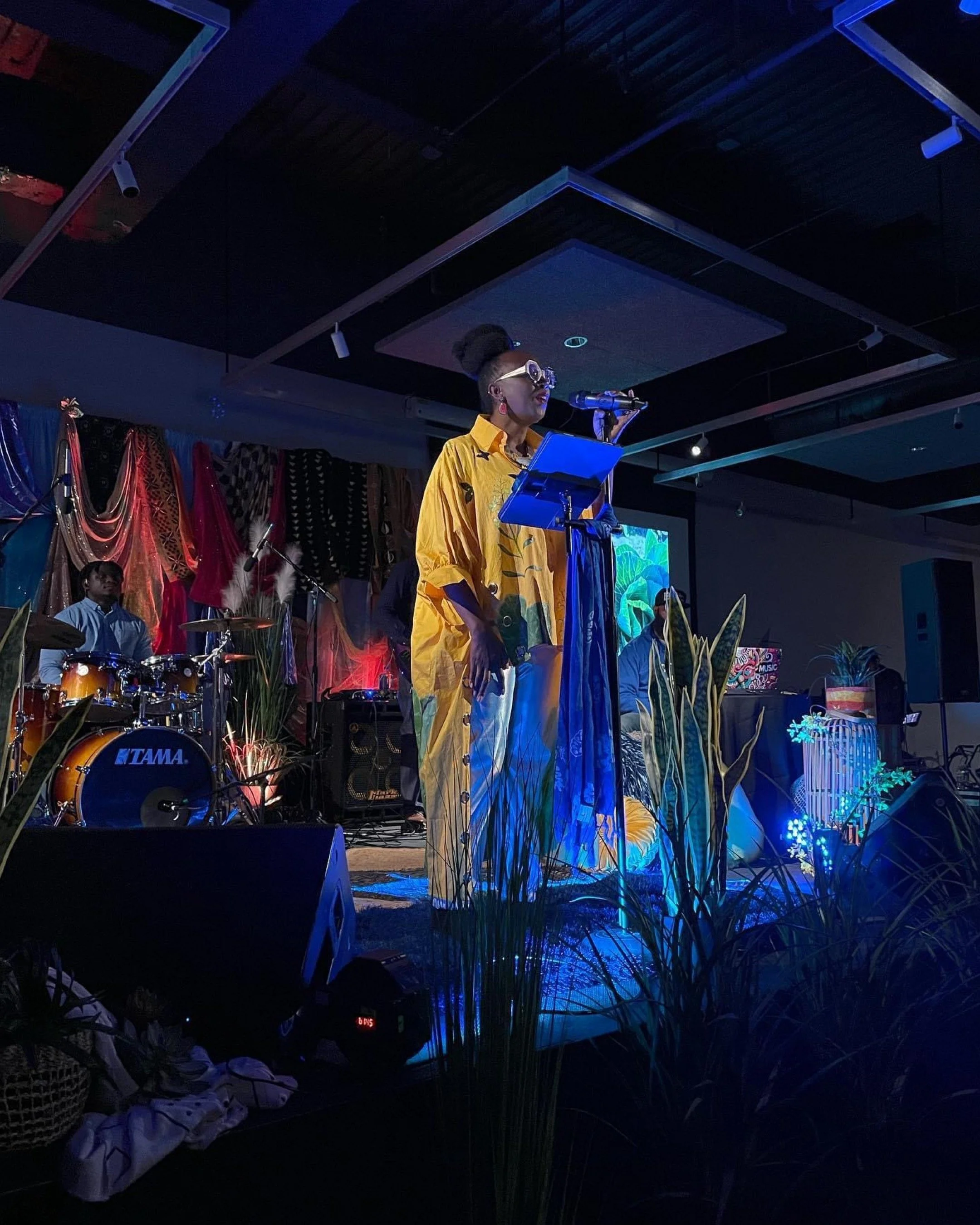 Mahoganee Amigér on stage, singing into a microphone. She is wearing a yellow dress and large white glasses. On stage there is also a drum kit and foliage.