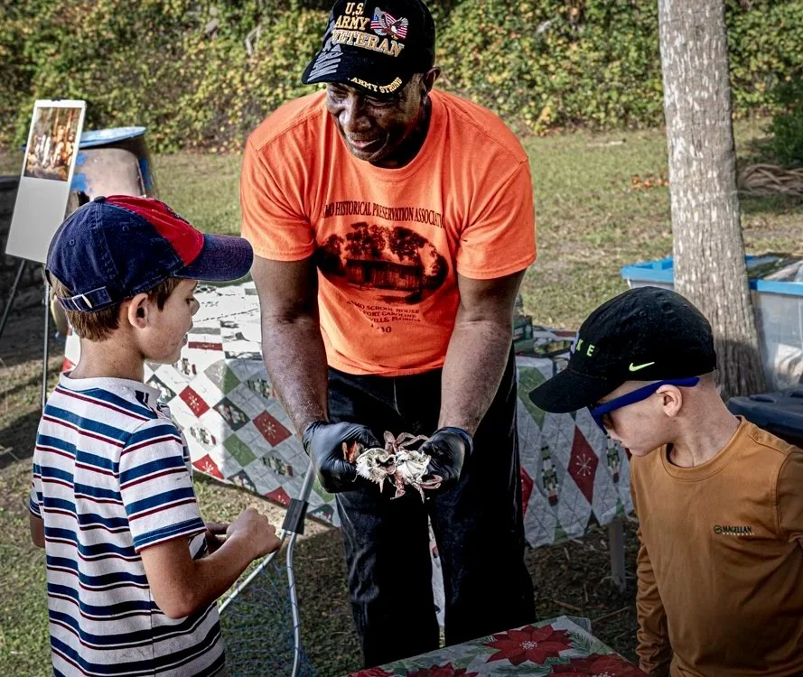 Leevon White, wearing an orange shirt, a US Army Veteran baseball cap, and black latex gloves holds a crab to demonstrate to two young boys.
