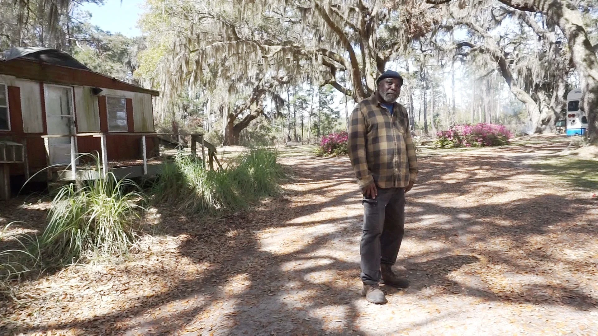 Stanley Walker wearing a beanie, a flannel shirt, and black jeans standing outside near a large willow tree. Pink flowers, grass, other trees, and a path through the garden in the background.