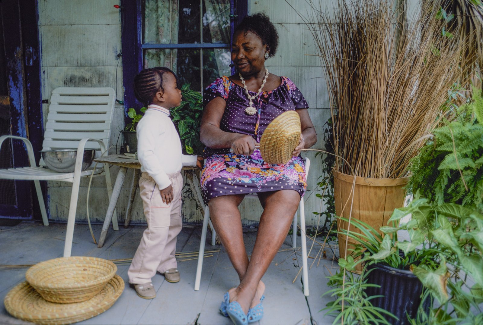 Historical image from 1987 of a Gullah Geechee woman sitting on a chair outside sewing a sweetgrass basket. A small child wearing white looks up at her and she smiles. In the background is another completed basket and a variety of foliage.