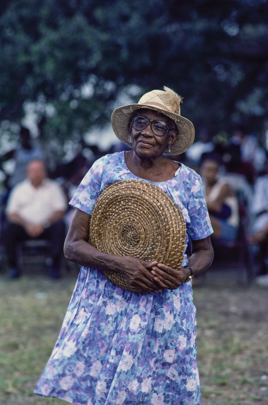 Historical image from 1987 of a Gullah Geechee woman wearing a purple floral dress, a sunhat with a feather, and large glasses carrying a sweetgrass basket.