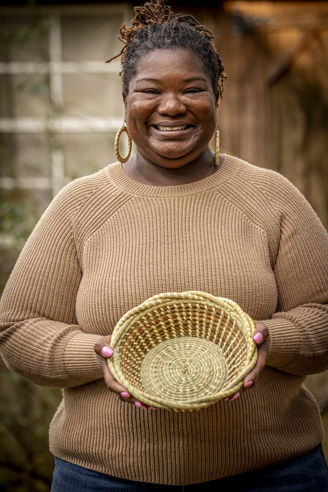 Andrea Cayetano Jefferson wearing a beige sweater and hoop earrings with pink painted nails. She is smiling and holding a sweetgrass basket.