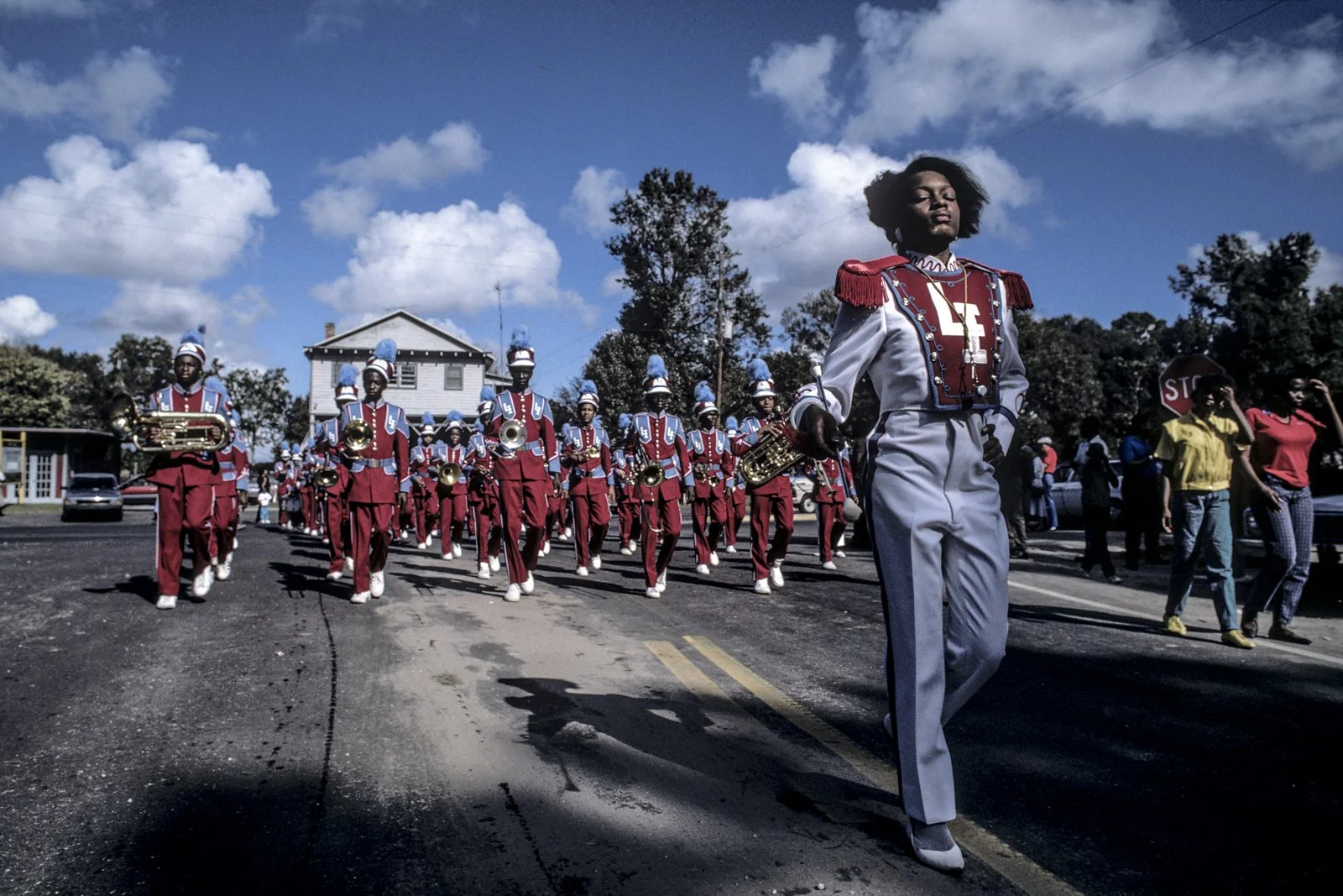 Historical image from 1987 of a marching band wearing blue and red. The person in the front is wearing a slightly different outfit and carrying a baton, while those in the background carry instruments.