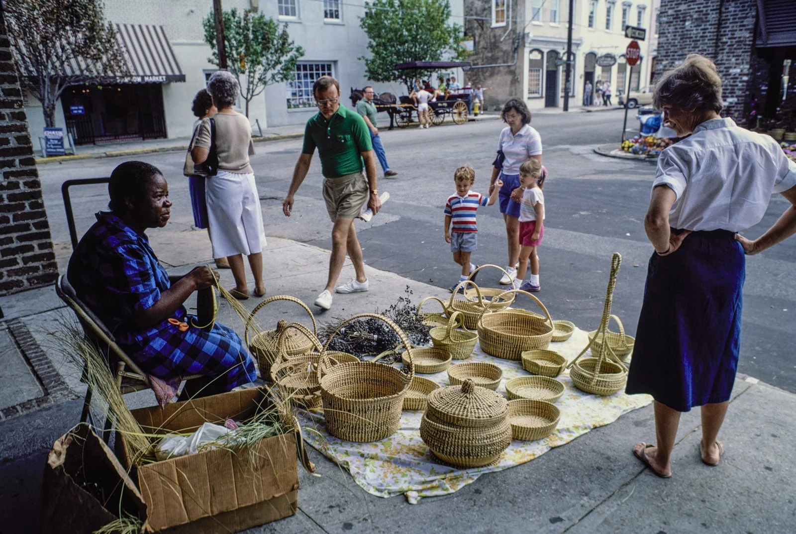 Historical image from 1987 of a Gullah Geechee woman sitting in a chair making sweetgrass baskets at a market. Completed baskets sit on a blanket in front of her. Families of white tourists appear to be walking up to her, looking at her work.