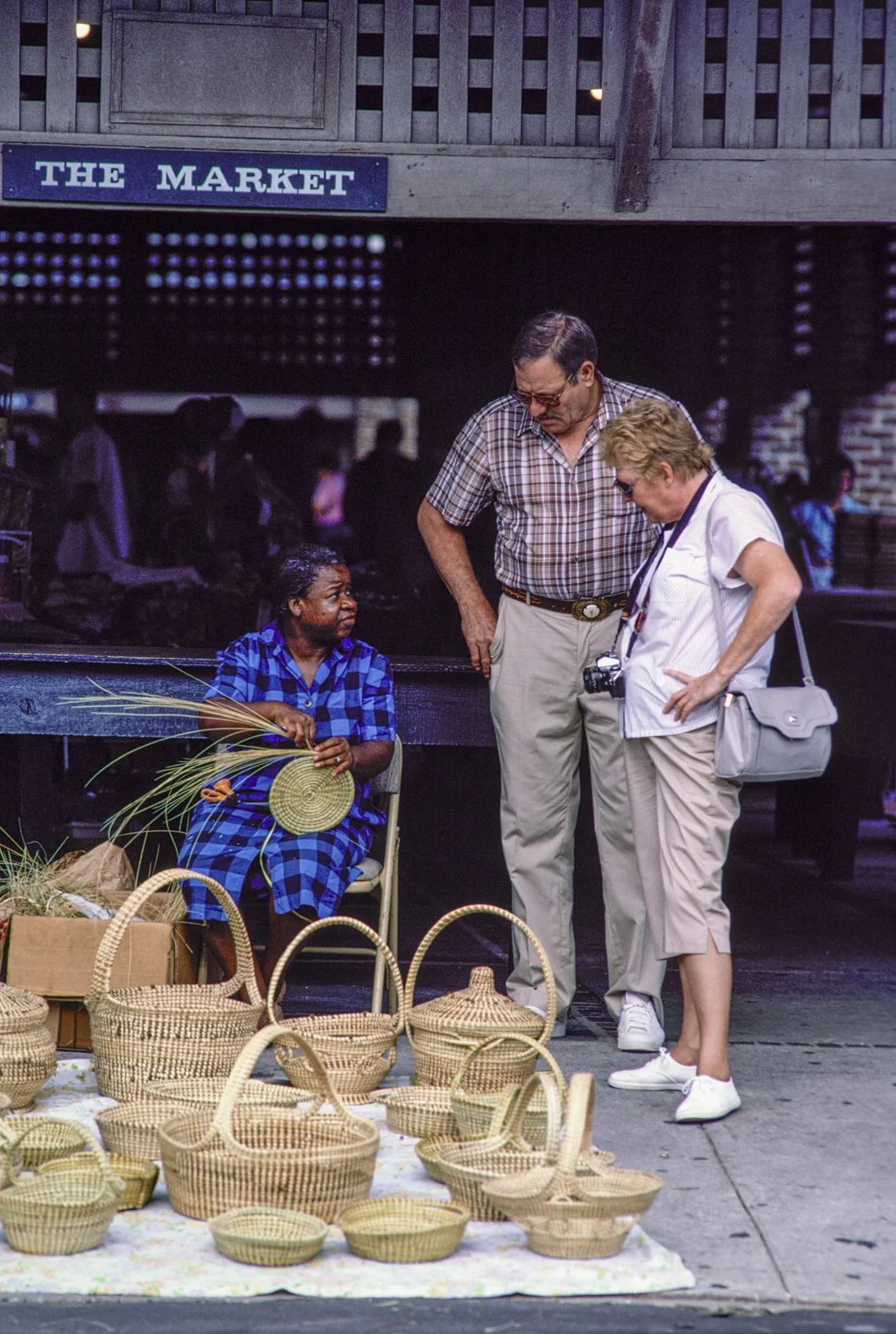 Historical image from 1987 of a Gullah Geechee woman sitting in a chair making a sweetgrass basket at a market. More baskets are set out in front of her. Two white tourists have stopped, hands on their hips, to speak to the woman.