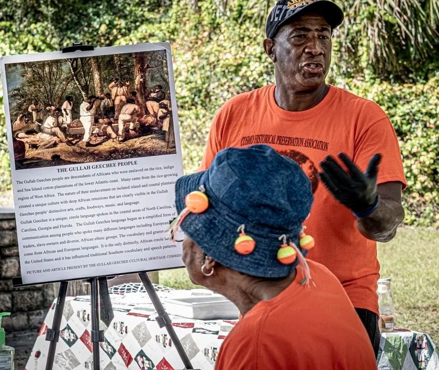 Leevon White and another woman are pictured wearing orange t-shirts that read "Cosmo Historical Preservation Association." Leevon White looks out at the crowd, speaking, while standing next to a sign that reads "The Gullah Geechee People."