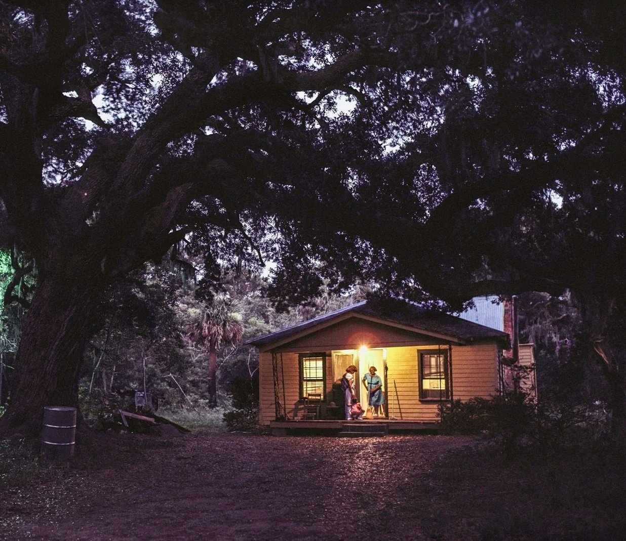 Historical image from 1987 of a house. It is dusk and the porch light is on. Two figures stand on the porch.