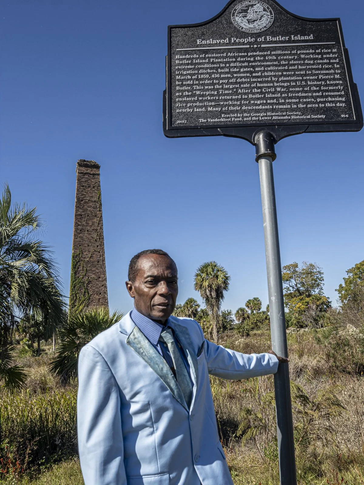 Griffin Lotson standing outside, touching a black sign that reads "Enslaved People of Butler Island." He is wearing a light blue suit. In the background is a brick monument, palm trees, and other foliage.