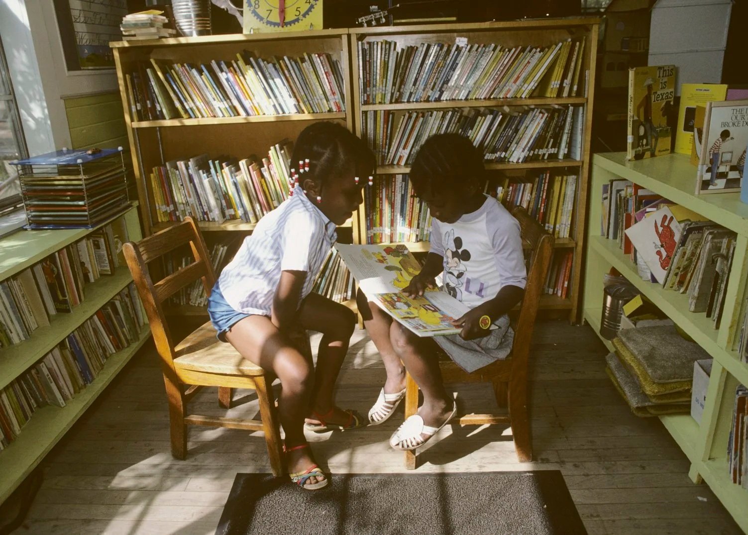 Historical image from 1987 of two young girls reading books together. The background contains shelves with many more children's books.