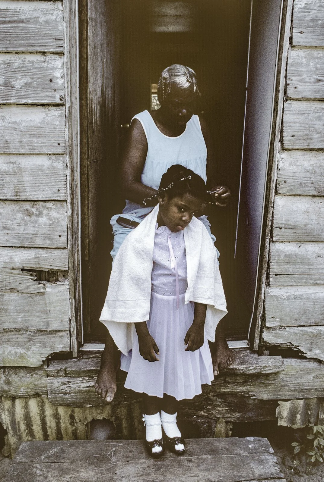 Historical image from 1987 of an older woman and a young girl standing outside. The girl is wearing a purple dress with a white towel draped across her shoulders, ad the older woman is standing behind her, doing her hair.