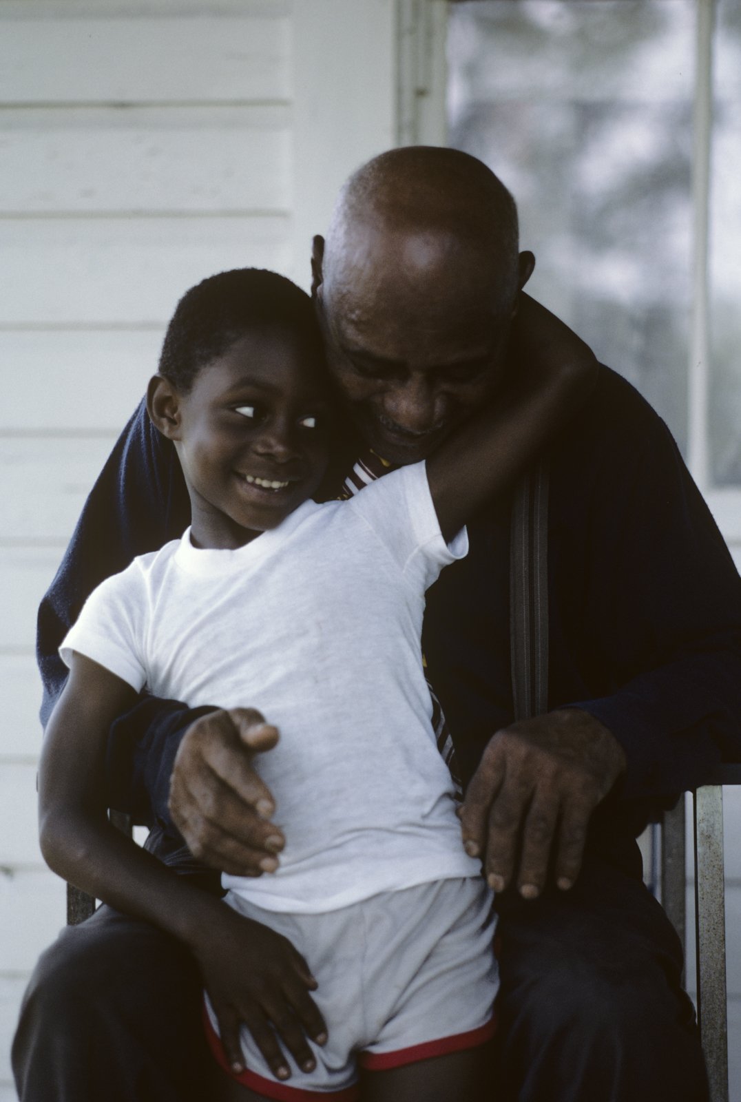 Historical image from 1987 of an older man and a young boy sitting together, smiling and hugging.