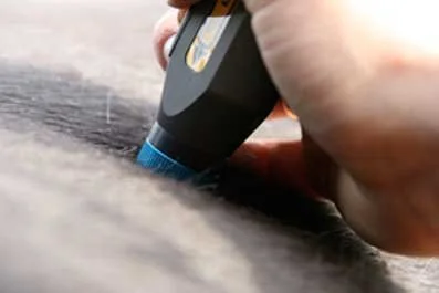 A hand using a black and blue tool to carve or etch on a dark surface.