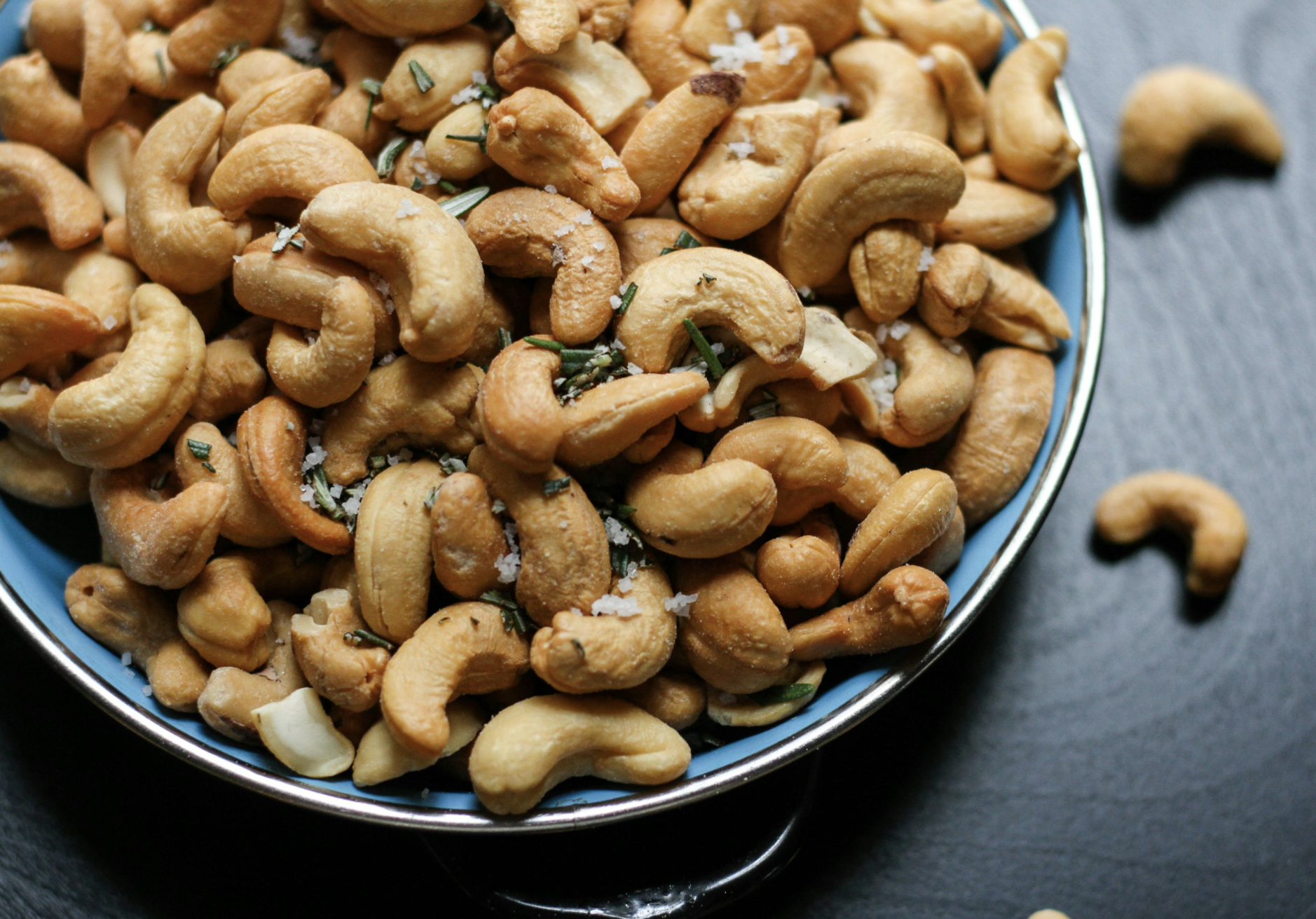 A bowl filled with roasted cashew nuts sprinkled with chopped herbs and coarse salt, with some cashews spilled on a dark surface beside the bowl.