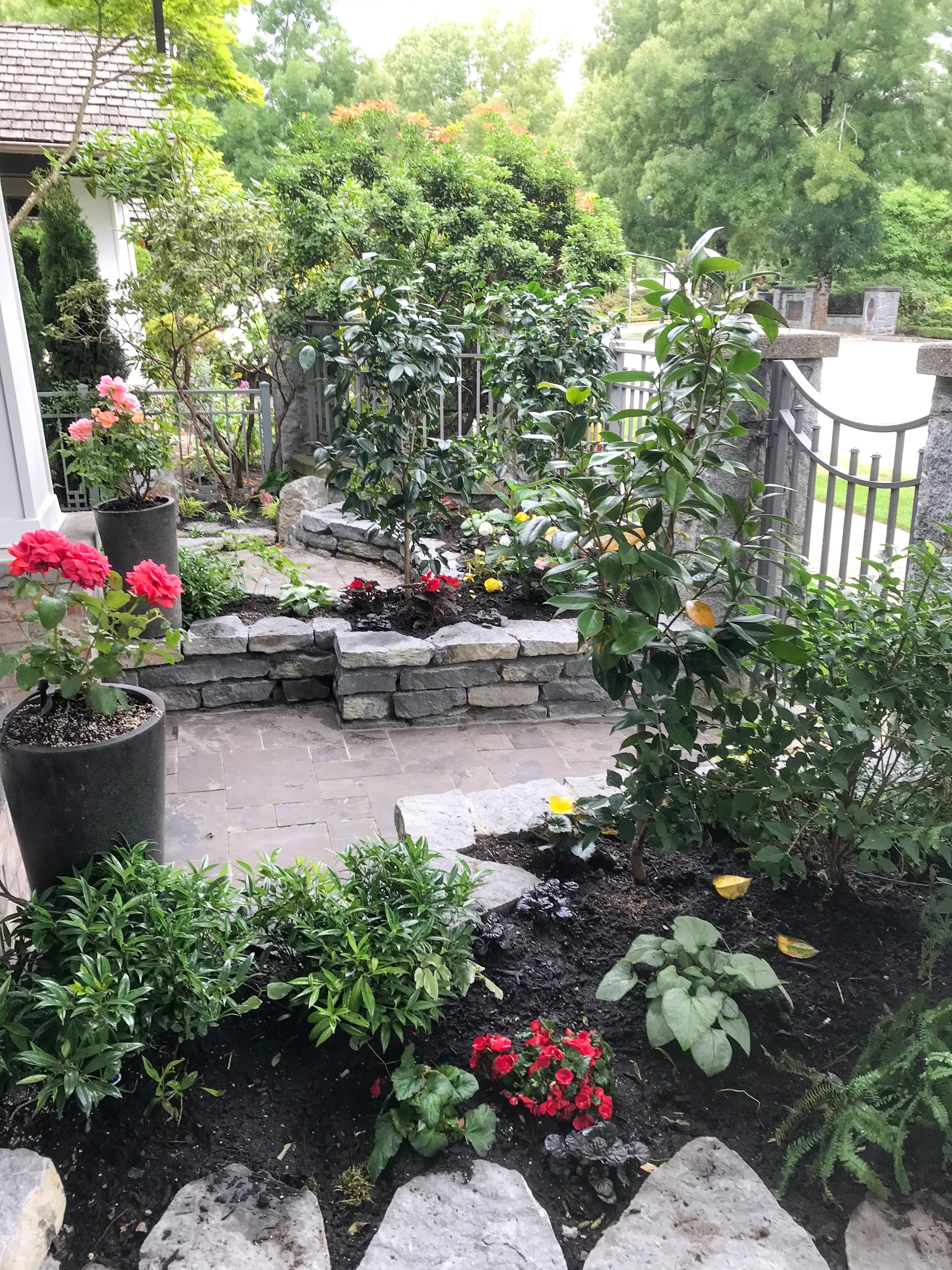 A lush garden with various flowering plants and green shrubs, bordered by a stone garden bed, and a metal fence with stone posts, set against a background of large trees and a house.