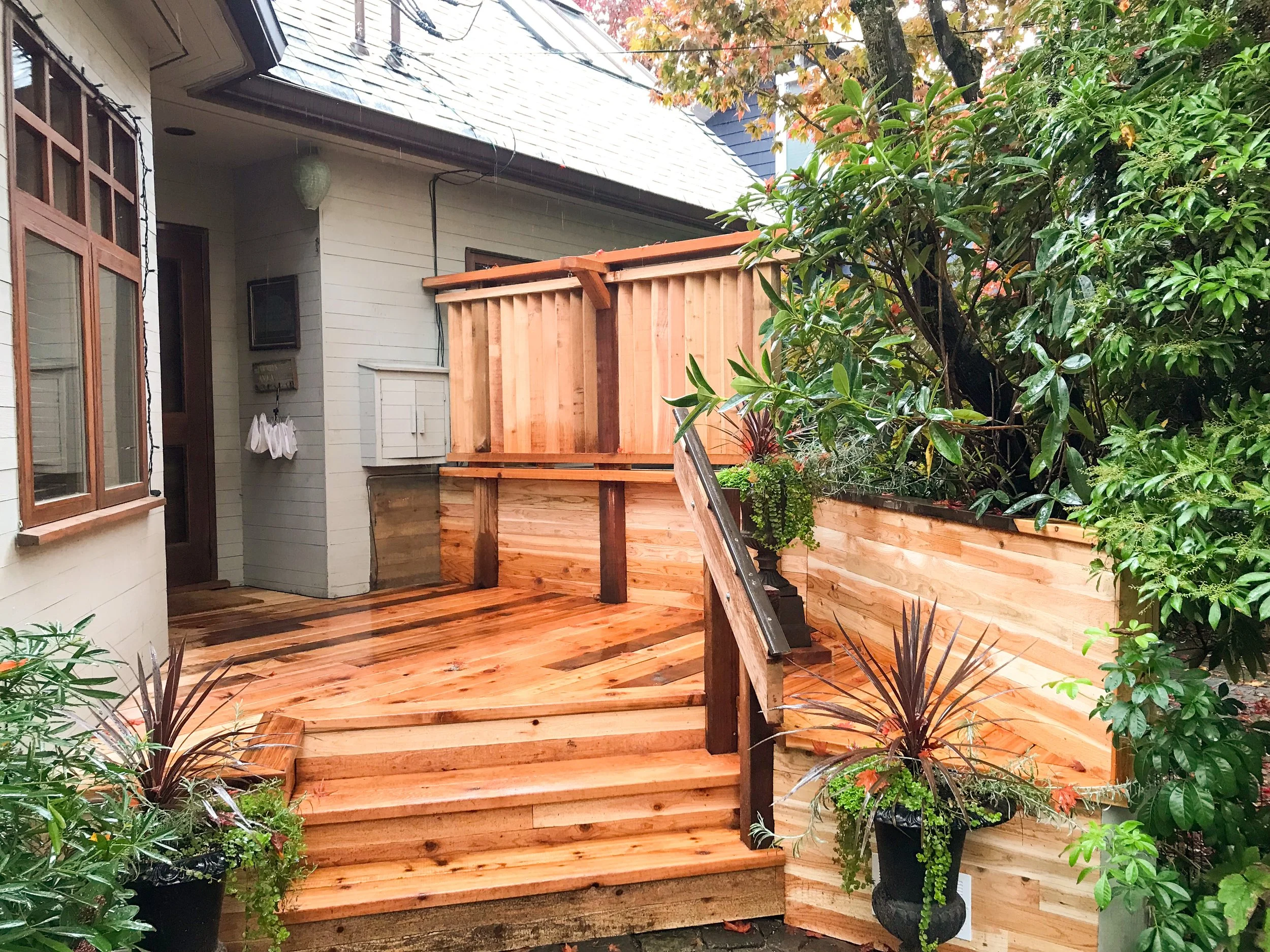 A newly constructed wooden deck with stairs attached to a house. The house has white siding, windows with wooden frames, and a gray roof. There is a green shrub with orange and red leaves in the background and potted plants on the deck.