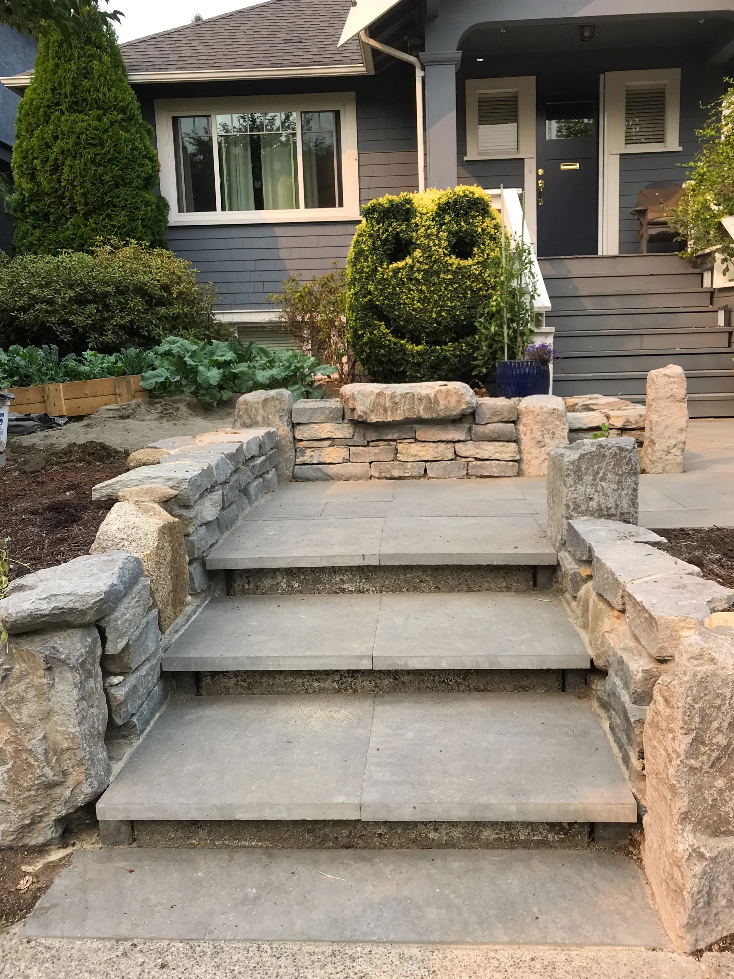 Concrete stairs leading up to a house entrance with stone wall borders on each side, surrounded by green bushes and plants.