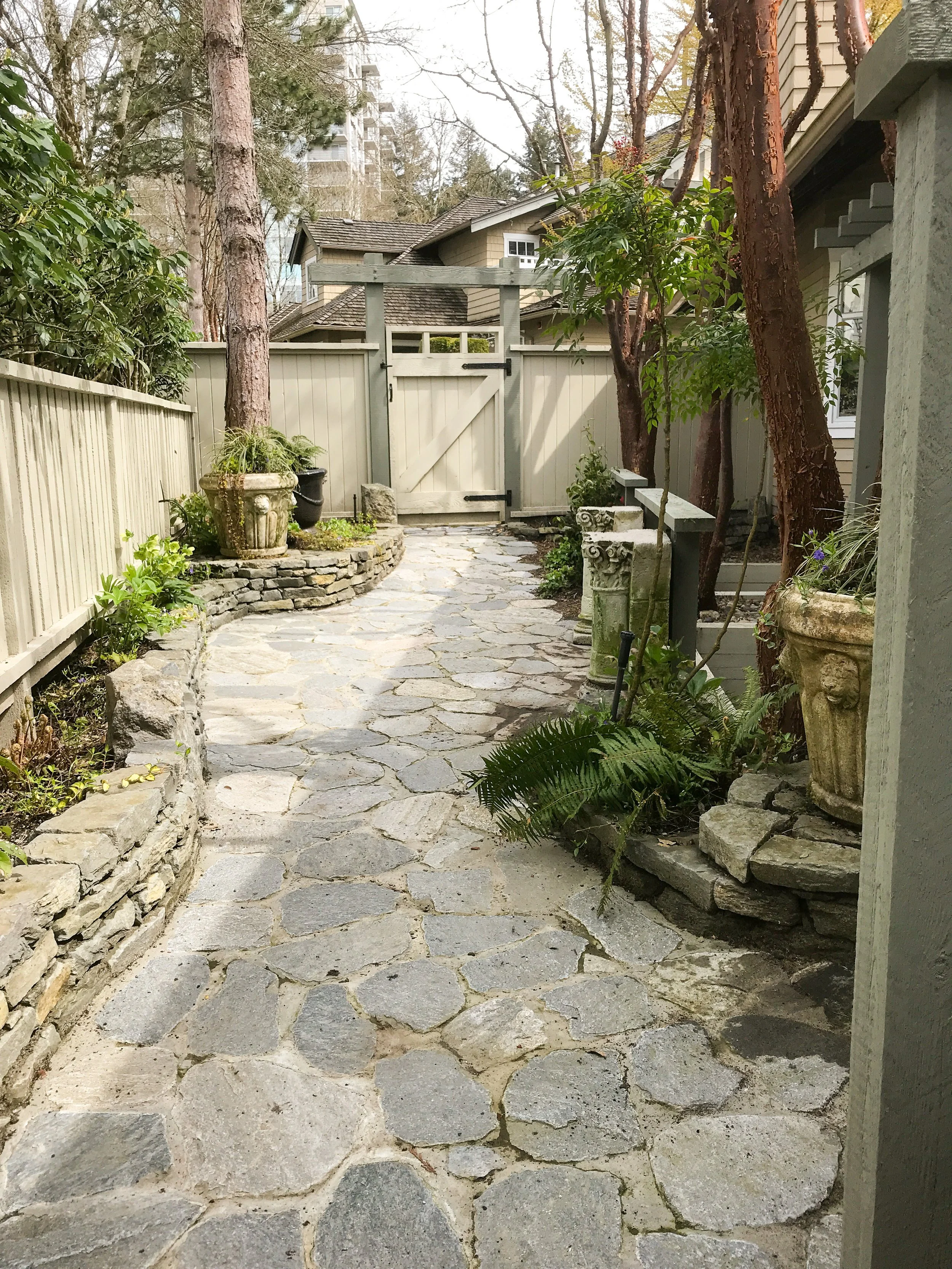 A stone pathway leading to a wooden gate, surrounded by potted plants, trees, and a beige house with a sloped roof in the background.