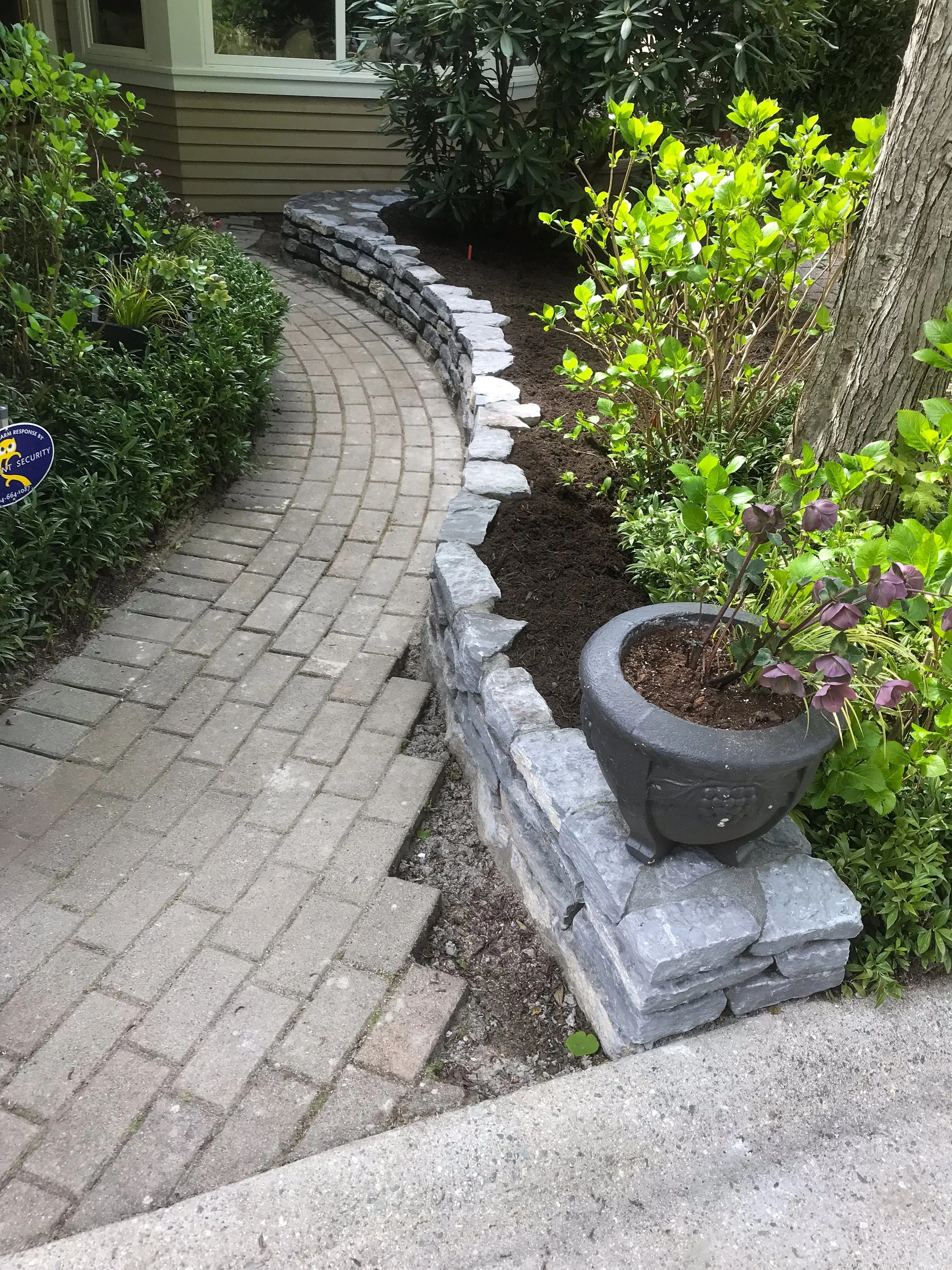 A curved brick pathway lined with gray stones and surrounded by green bushes and plants, including a large tree on the right side of the image and a house with horizontal siding and a window in the background.
