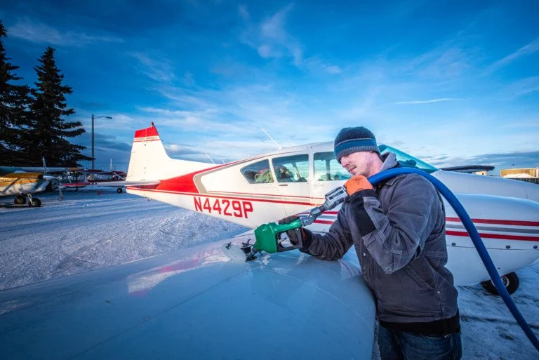 A man in winter clothing fills a small airplane with fuel at an outdoor airfield, snowy ground, blue sky, other planes and trees in the background.