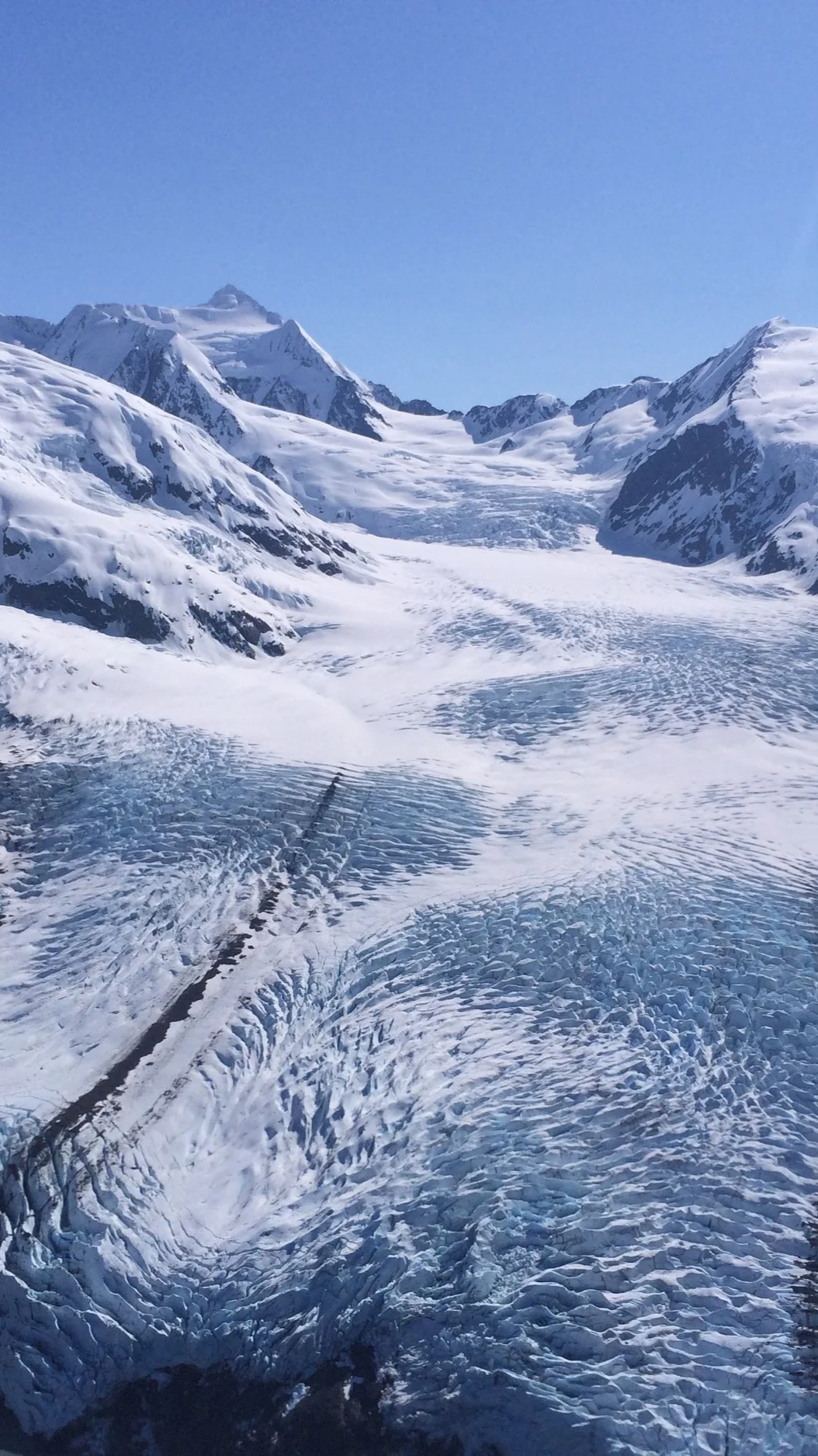 Snow-covered mountains and glaciers under a clear blue sky, with visible crevasses and mountain peaks in the distance. Flying in Alaska.