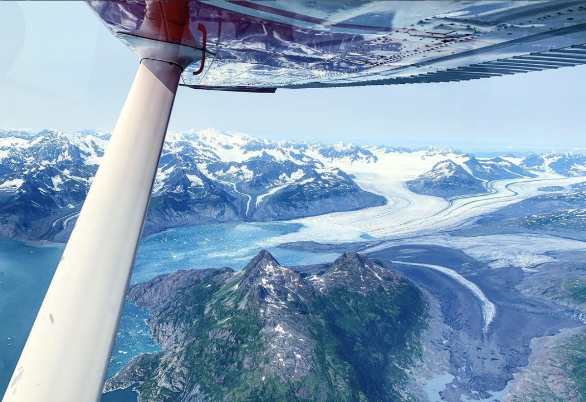 Aerial view from an airplane showing snow-capped mountains, glaciers, and a mountain with green and rocky terrain below.