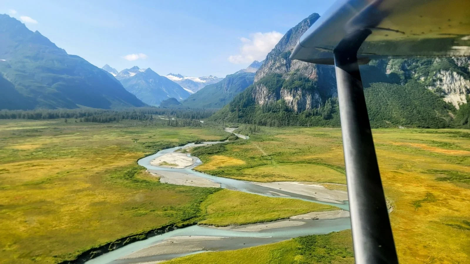 Aerial view of a winding river flowing through greenery with mountains in the background under a clear blue sky.